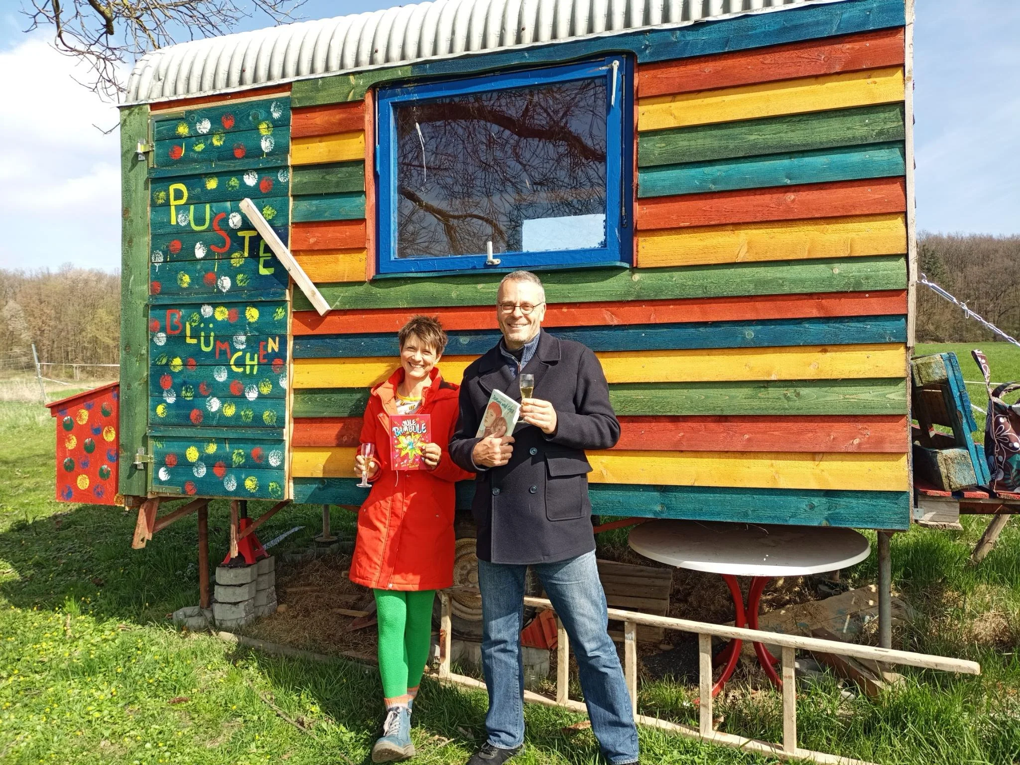 Judith und Andreas stehen mit ihrem Gut Pusteblume-Büchern vor einem bunten Bauwagen, lachen und haben Sektgläser in den Händen