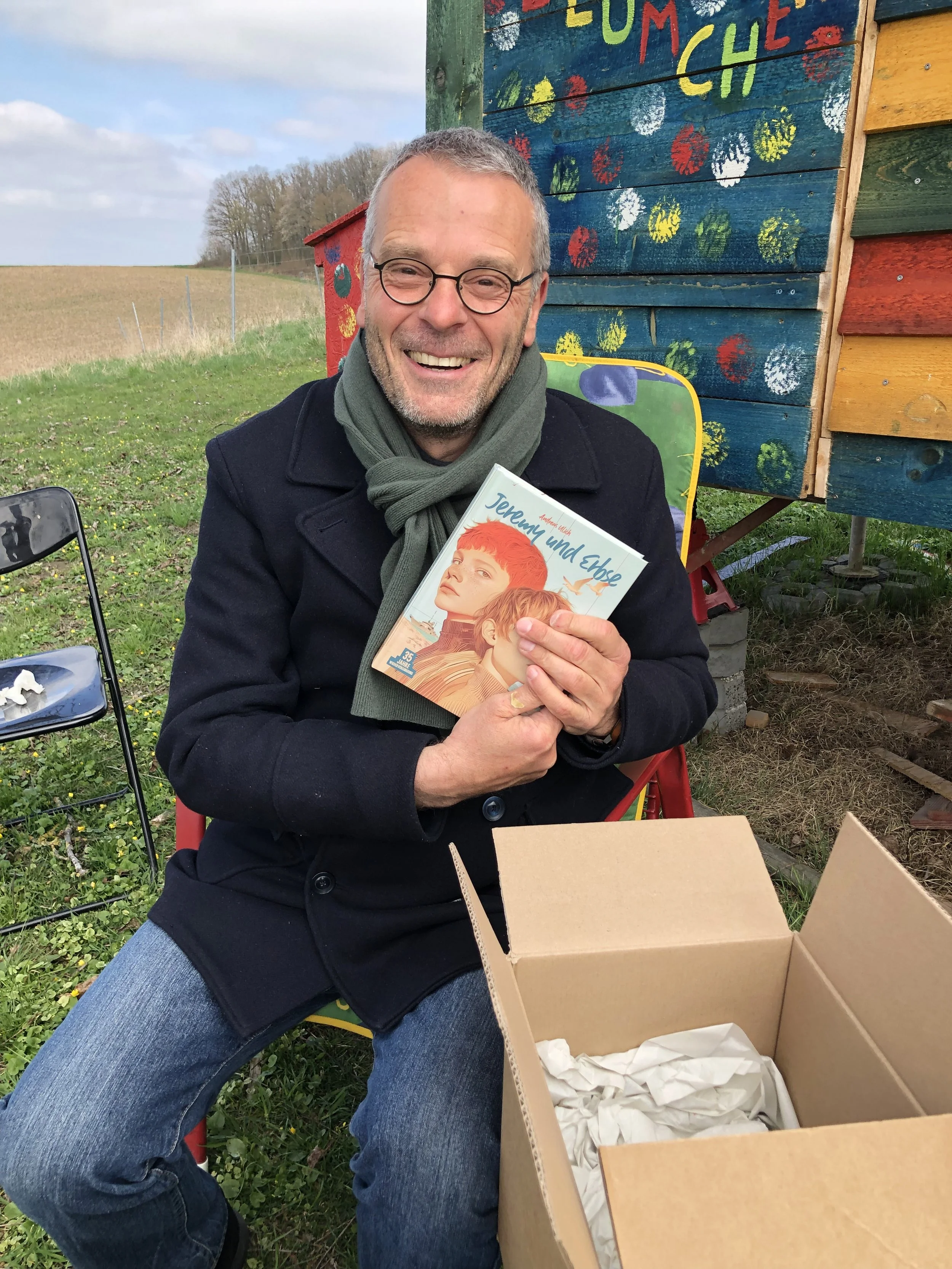 Andreas Ulich sitzt lachend im Garten von Gut Pusteblume und hält sein Kinderbuch Jeremy und Erbse in den Händen. Neben ihm steht ein offener Karton mit weiteren Büchern. Im Hintergrund ist ein bunt bemaltes Holzhaus zu sehen.