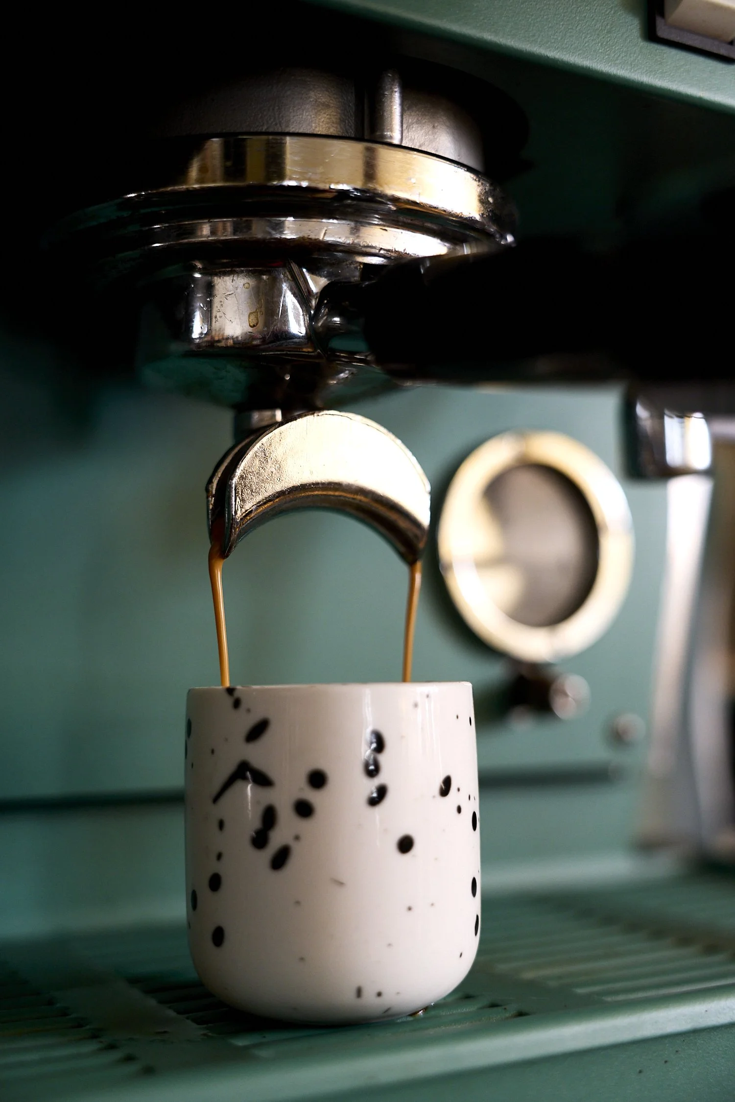 Close-up of a coffee or espresso machine dispensing dark coffee into a white cup with black specks, set on a green kitchen countertop.