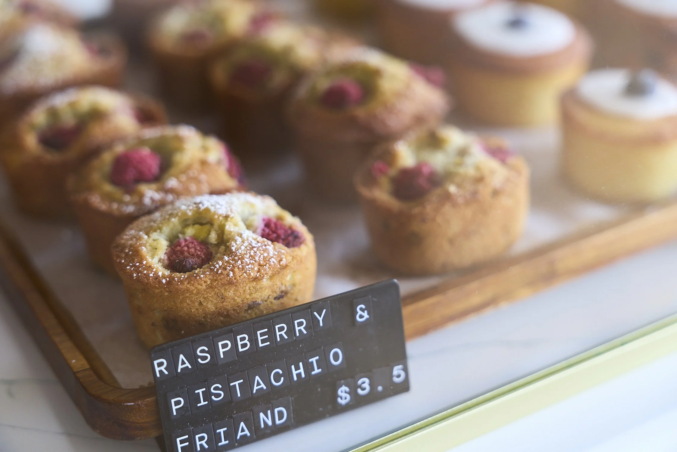 Close-up of raspberry and pistachio friands on a wooden tray with a black tag displaying ingredients and price.