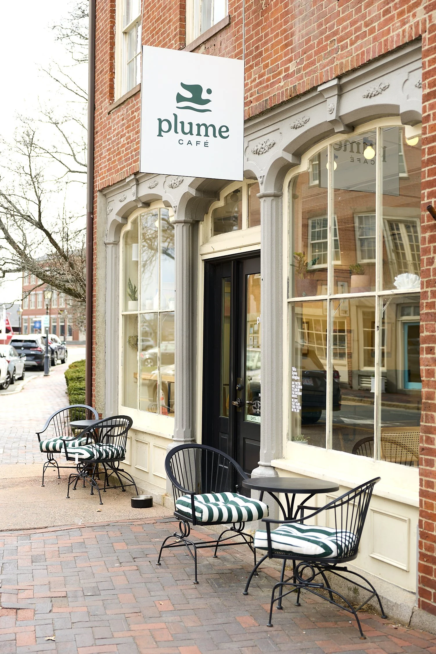 The exterior of a café called 'Plume Café' with outdoor seating. There are black metal chairs with striped cushions, and a small round table on a brick sidewalk in front of large windows with decorative framing.