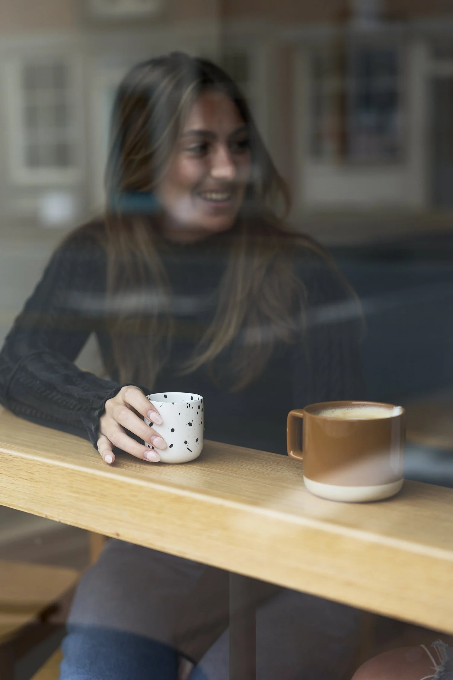 A smiling young woman with long brown hair, wearing a black long-sleeve top, holding a white mug, sitting at a wooden table with another brown mug nearby, seen through a window.