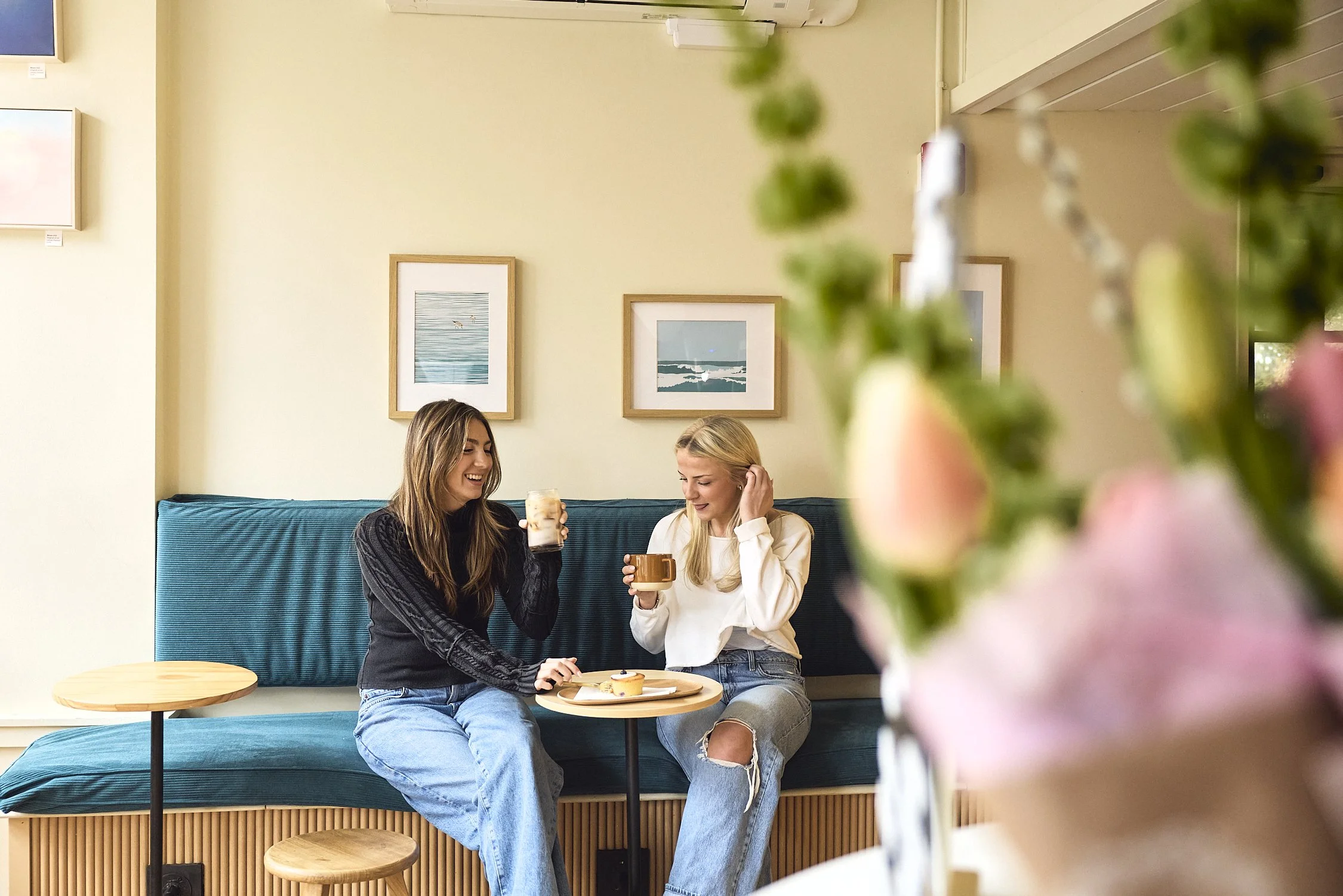 Two women sitting on a blue cushioned bench in a cafe, sharing a moment of laughter while holding drinks. One woman is dressed in black with long hair, the other in white with a ripped jeans. The scene features framed artwork on the wall behind them and floral decorations in the foreground.