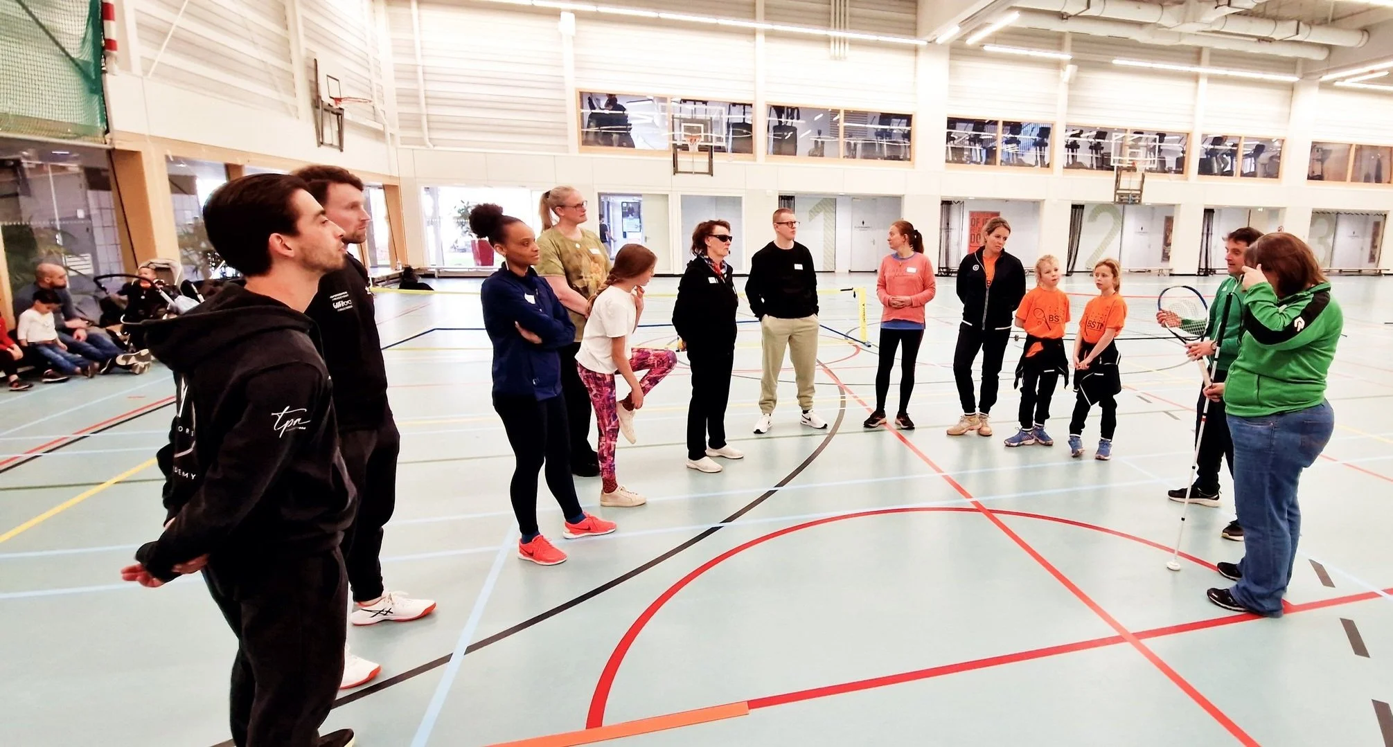 Foto van groep mensen die in halve cirkel staat. in een sporthal met blauwe vloer.