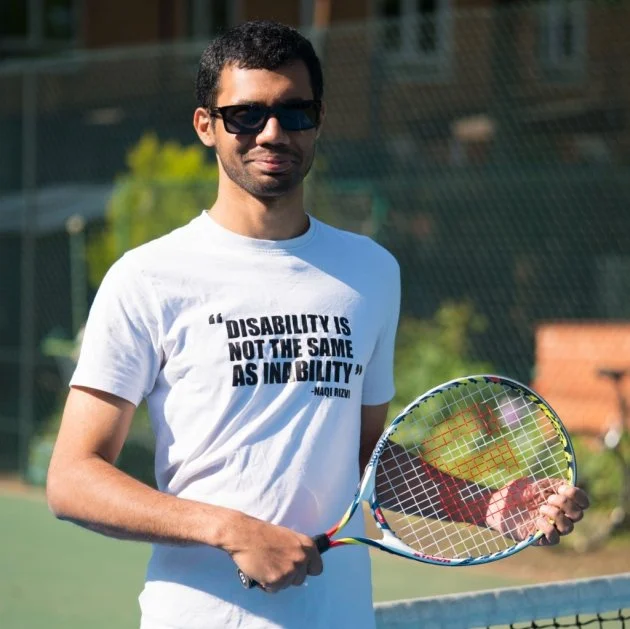 Portret van Naqi met zonnebril op en tennisracket in de hand. Op zijn witte t-shirt staat met grote zwarte letters: disability is not the same as inability.
