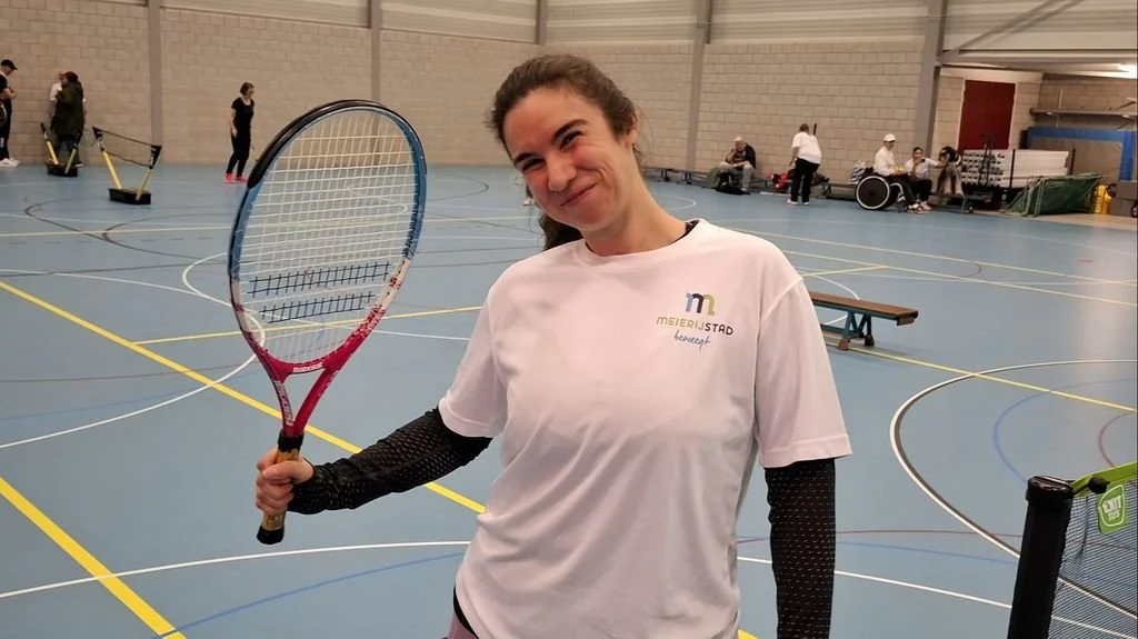 Vrouw lacht met tennisracket in de hand in een sporthal.