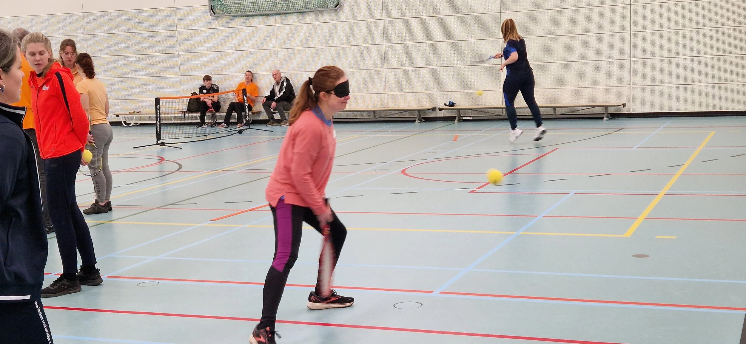 Vrouw staat met tennisracket klaar om de bal te ontvangen. De bal zweeft in de lucht een paar meter voor haar. Ze staat in een sporthal met een blauwe vloer. 