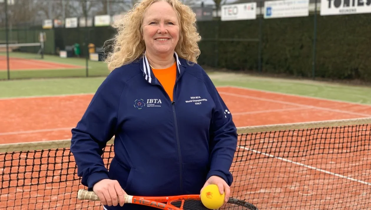 Fanny staat op de tennisbaan in haar sportkleren. Ze heeft een tennisracket en tennisbal in haar handen. De wind lijkt zachtjes door haar haren te waaien.