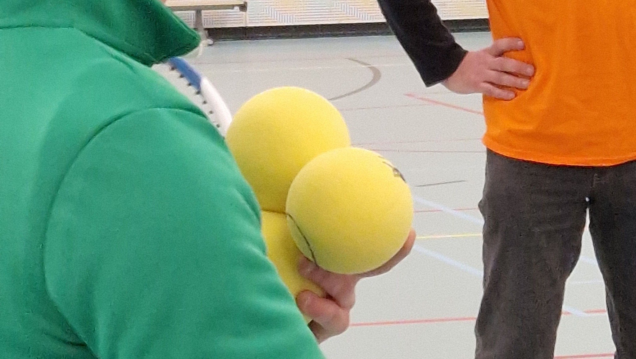 Close-up van drie ballen in de hand. Schouder  ook beeld met groen-blauw t-shirt. Op de achtergrond staat iemand met een zwarte broek en oranje t-shirt. Tot borsthoogte in beeld. 