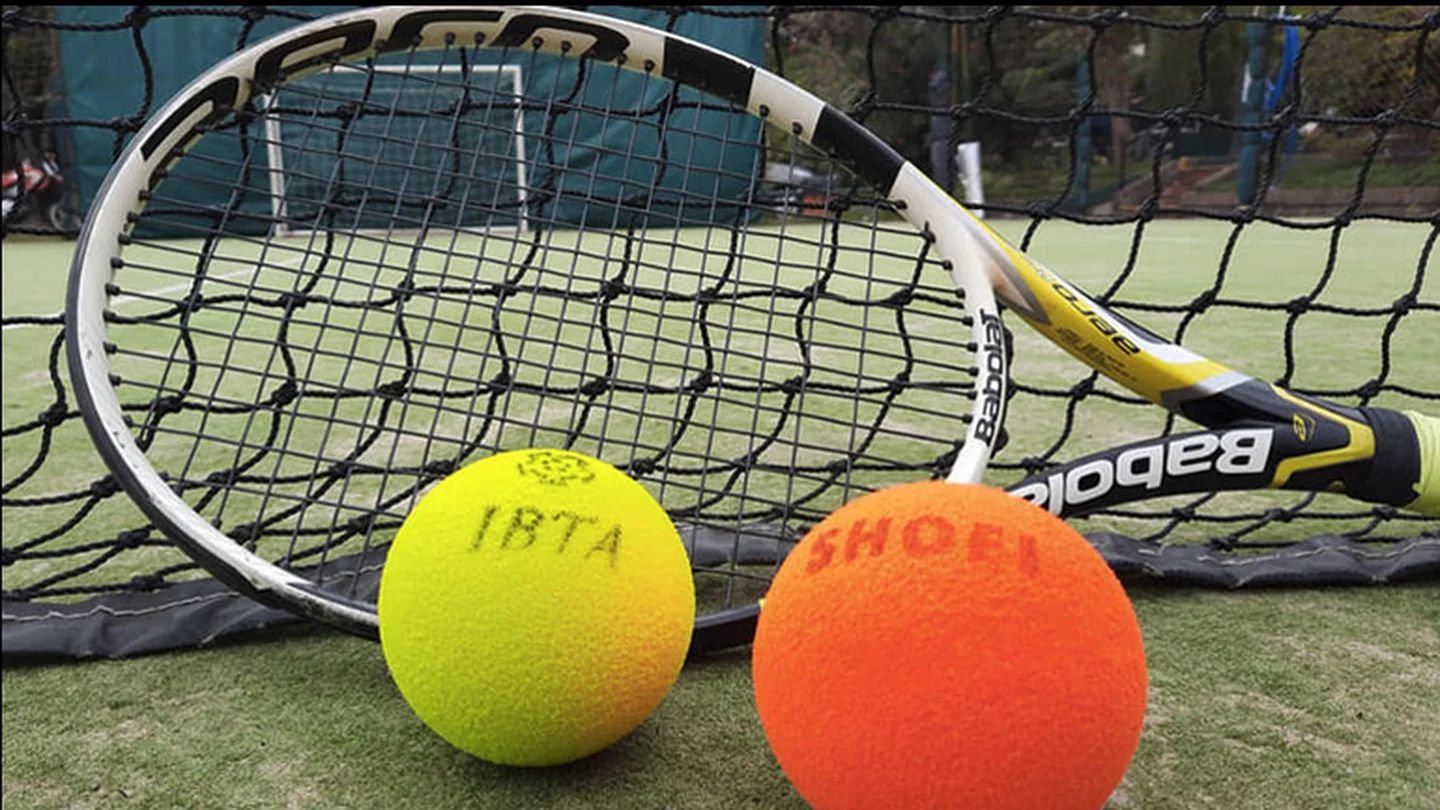 Tennisracket staat tegen net aan. Voor het tennisracket liggen twee tennisballen: een gele en een oranje. 