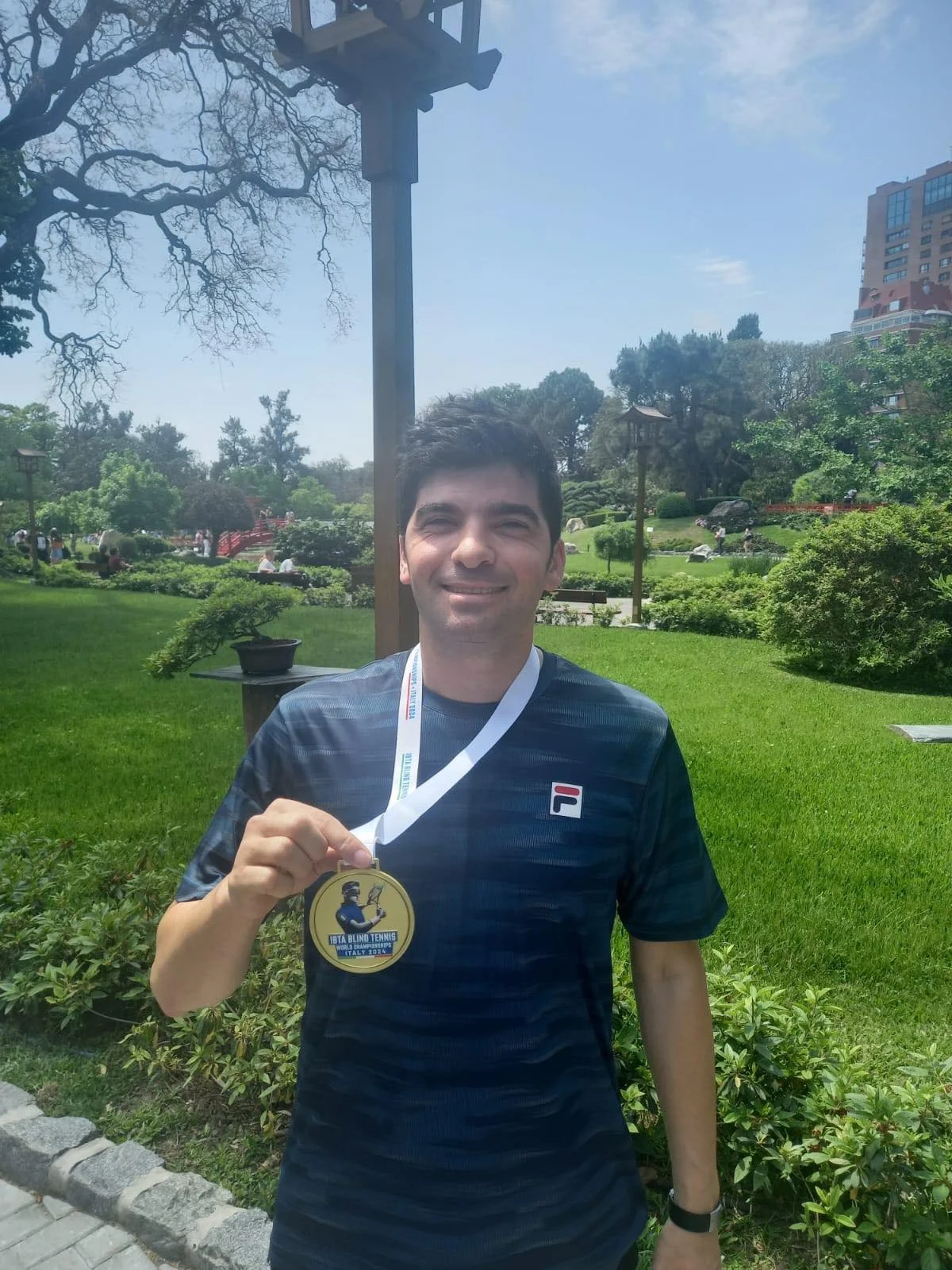 Portret van een lachende Naqi met  een mediaille in op zijn nek die hij uitsteekt naar de camera met zijn hand. Hij staat in een groene omgeving met blauwe lucht