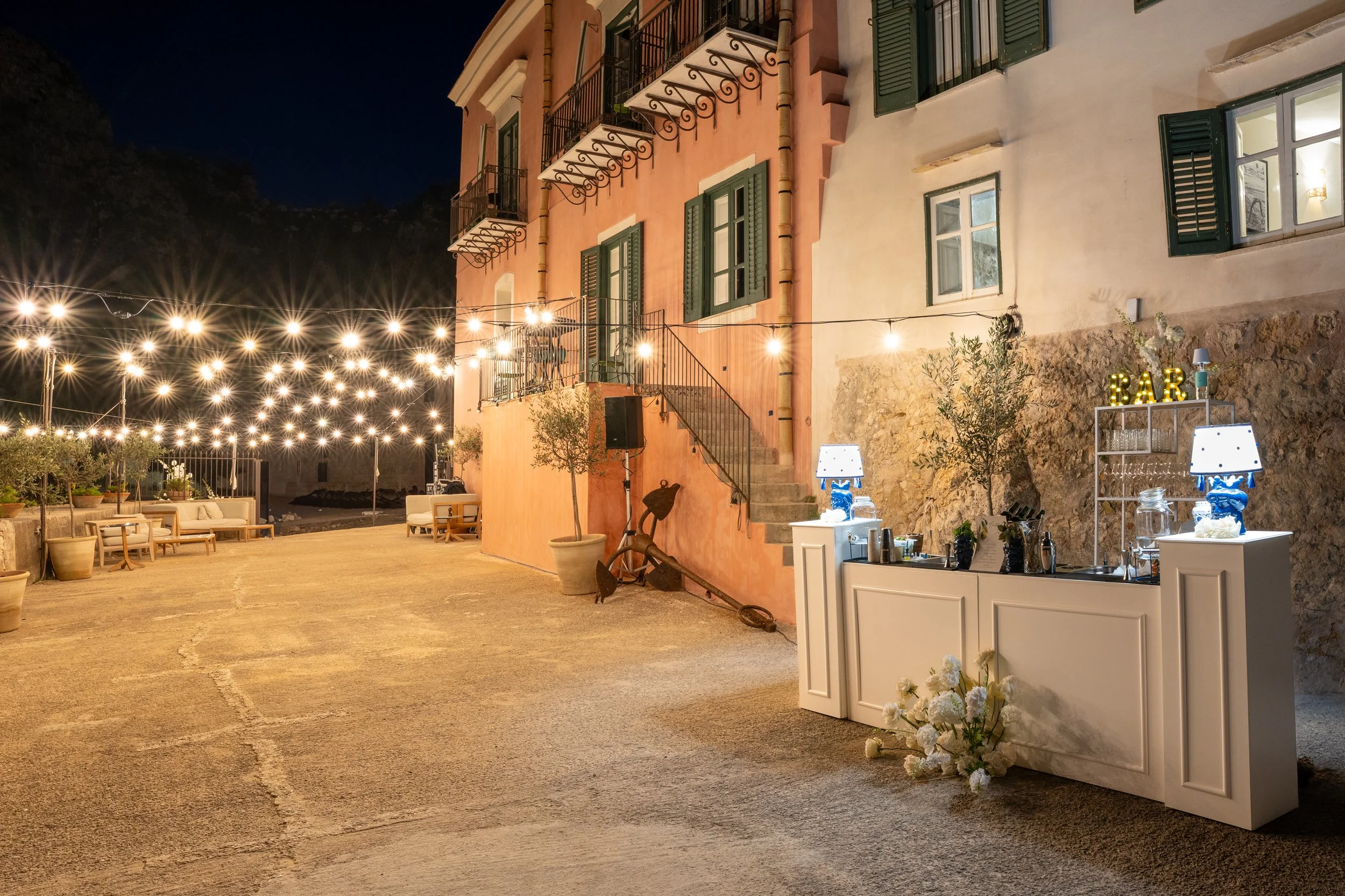 Vicolo di sera con luci decorative appese, un bar all'aperto, piante in vasi e un edificio con balconi e finestre aperte.