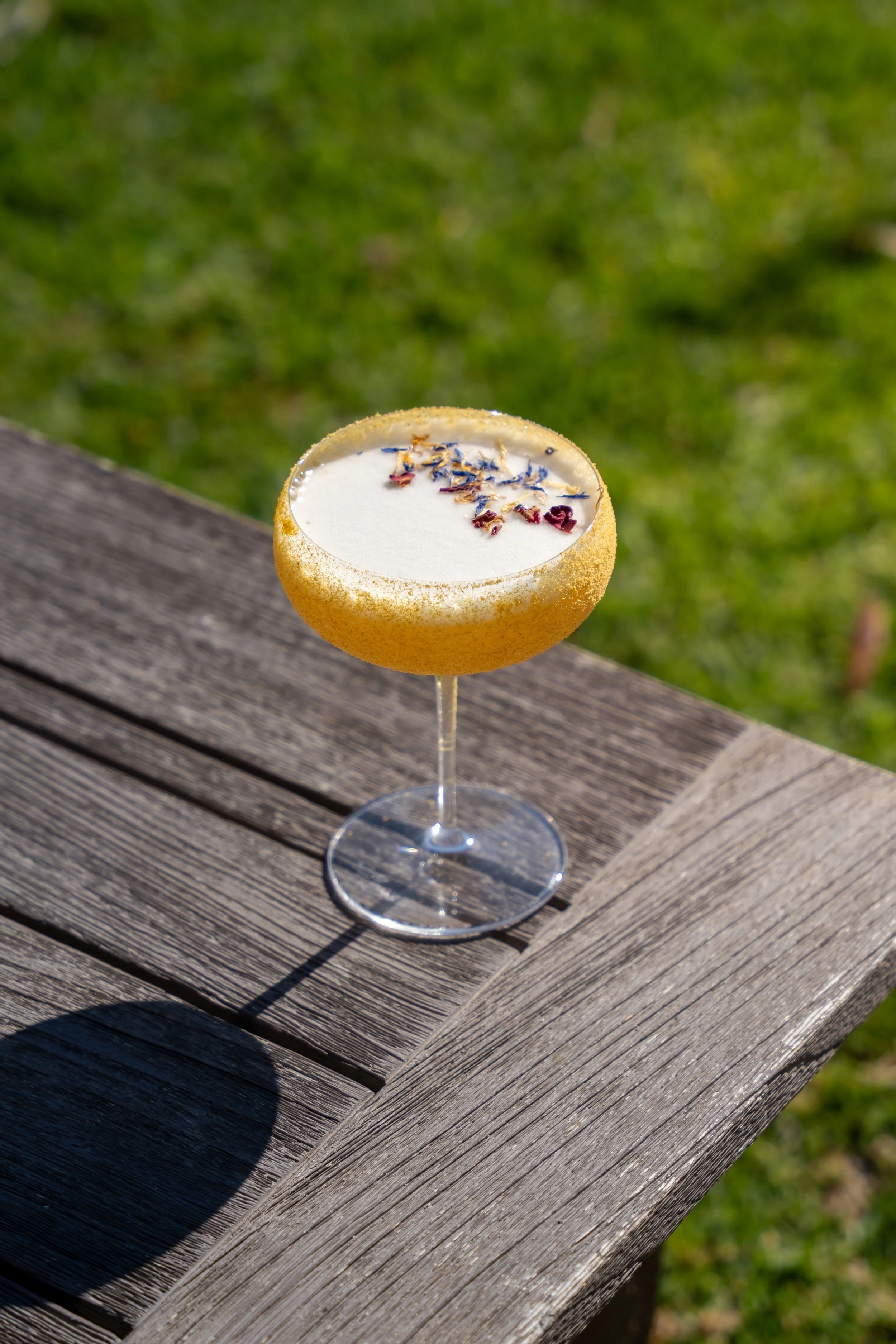 Cocktail in una coppa con bordo zuccherato, decorato con fiori secchi, su un tavolo di legno all'aperto.