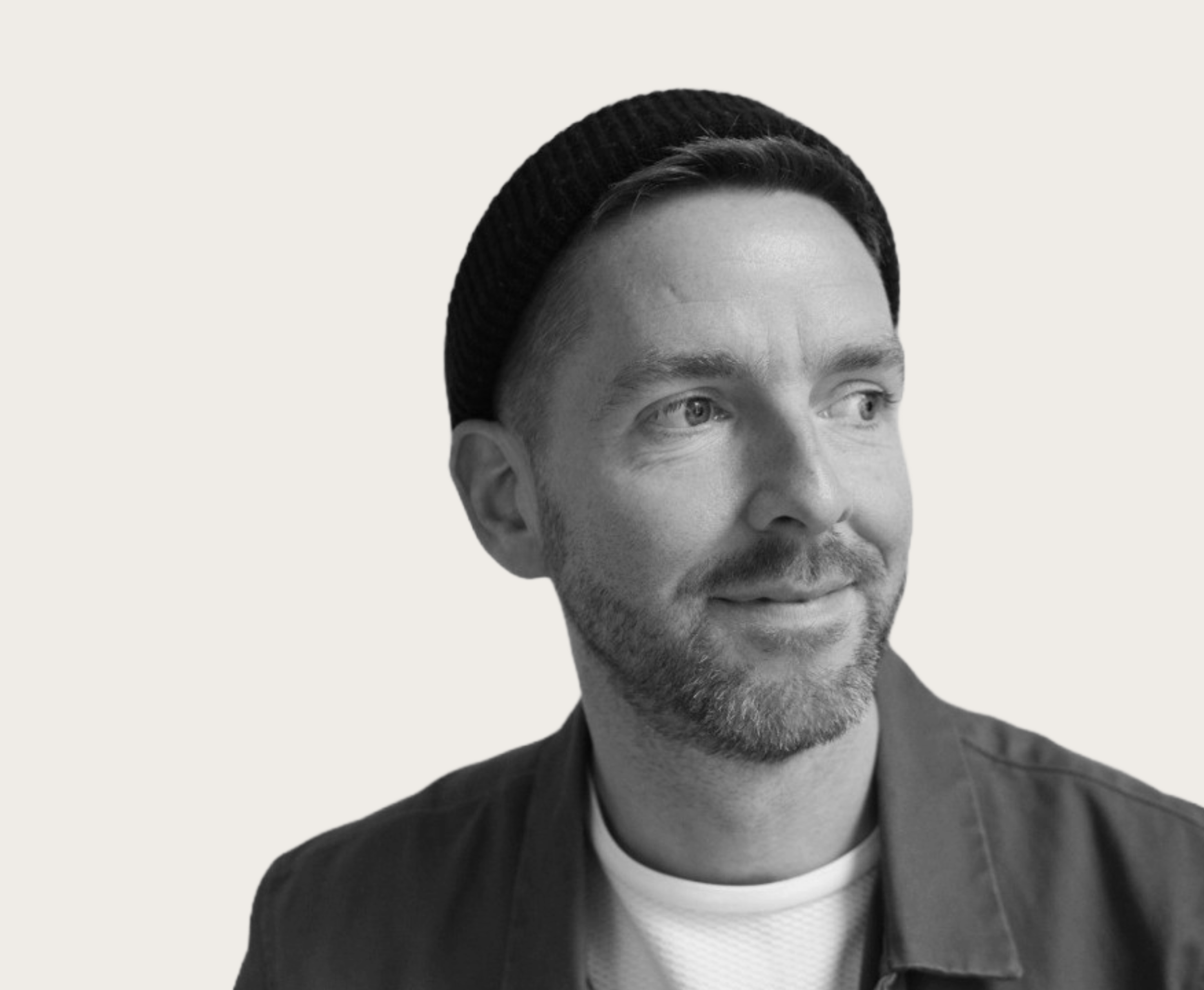 Black and white portrait of a man wearing a beanie and jacket, looking to the side and smiling.