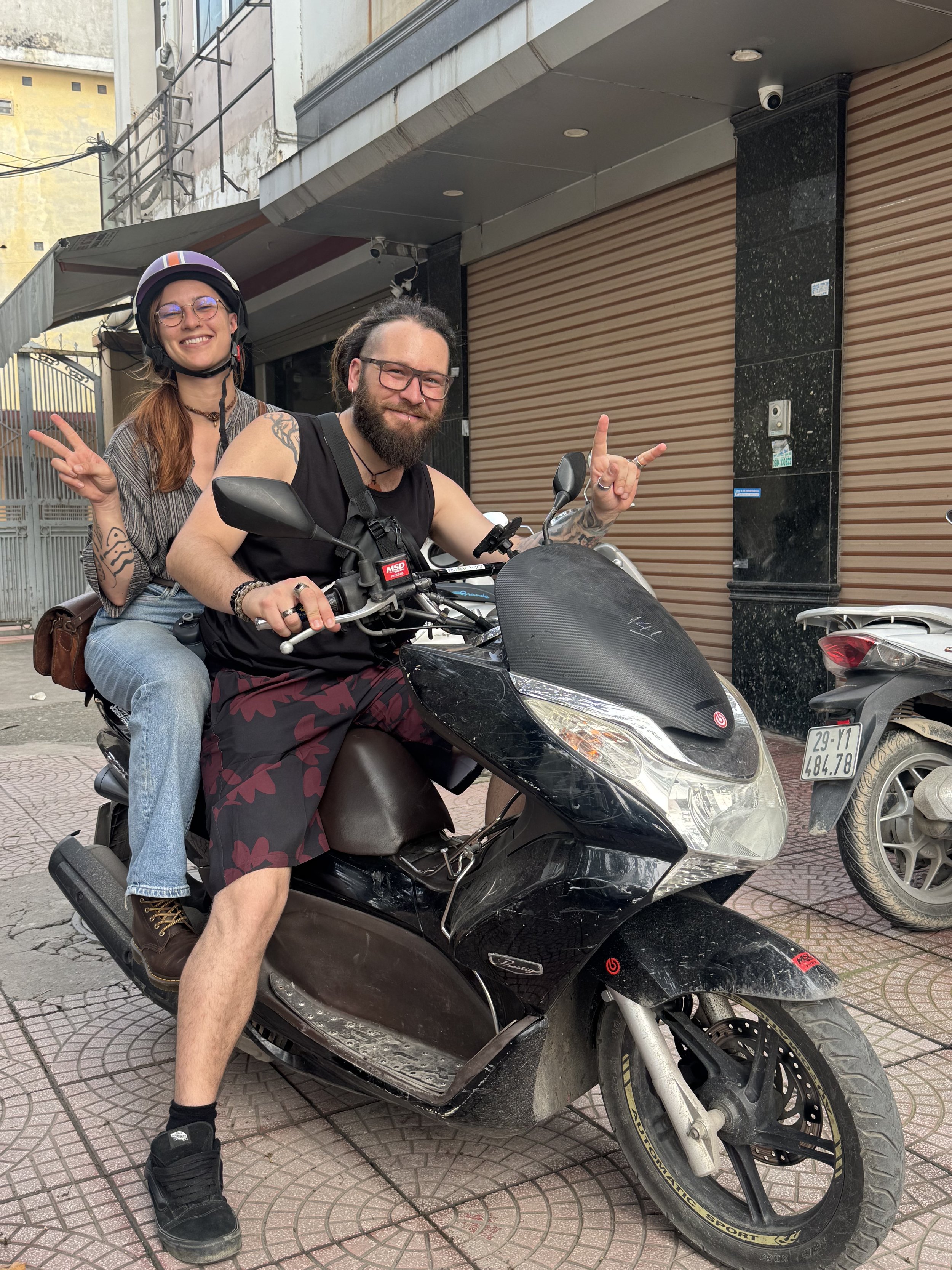 Two people sitting on a parked motorcycle, one wearing a helmet, both smiling and making peace signs, on a city street with shops in the background.