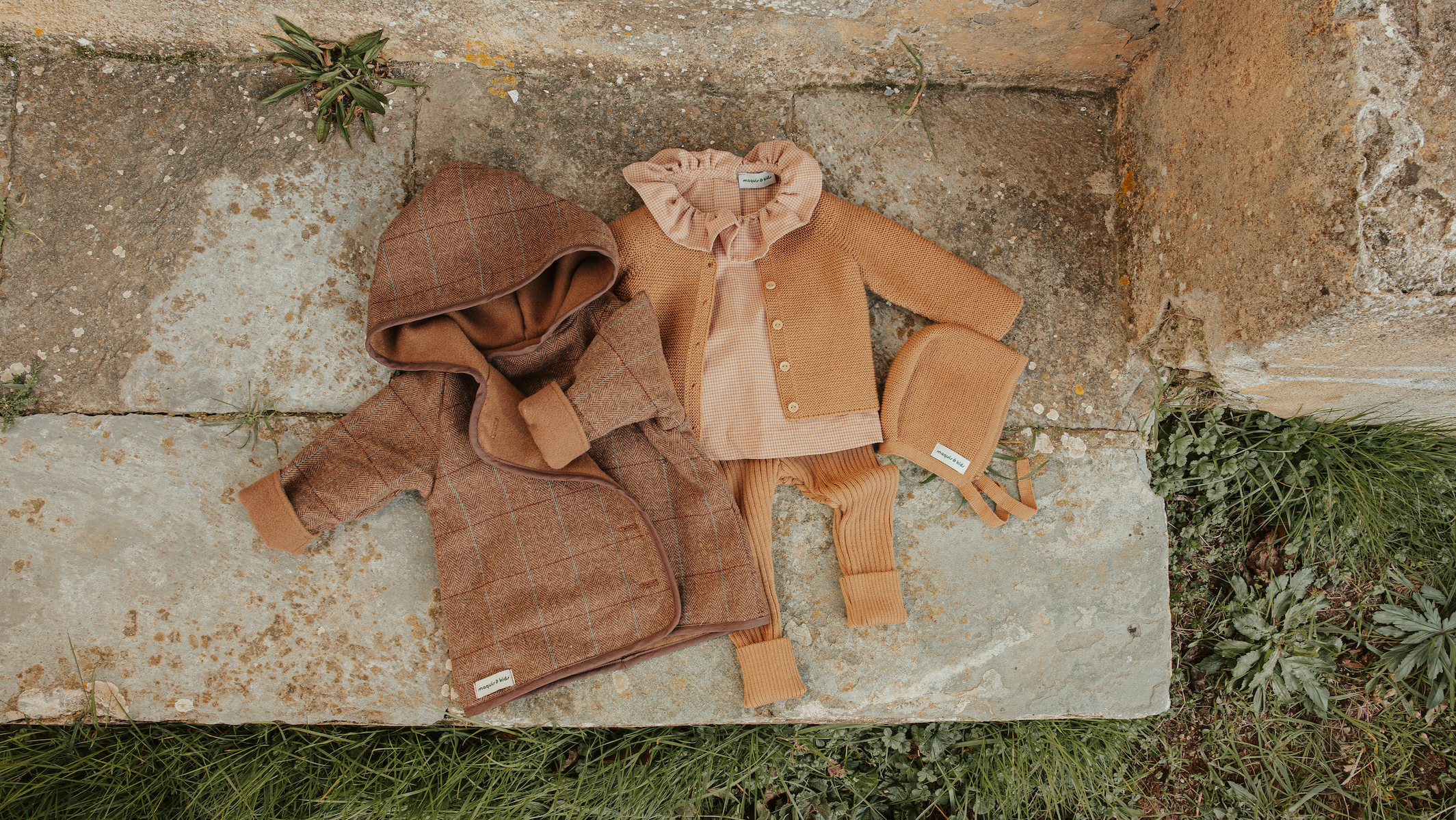 Vêtements pour enfants en tons marron et beige disposés sur un sol en pierre avec des plantes autour, comprenant un manteau, un cardigan, un chapeau, une écharpe et une cagoule.