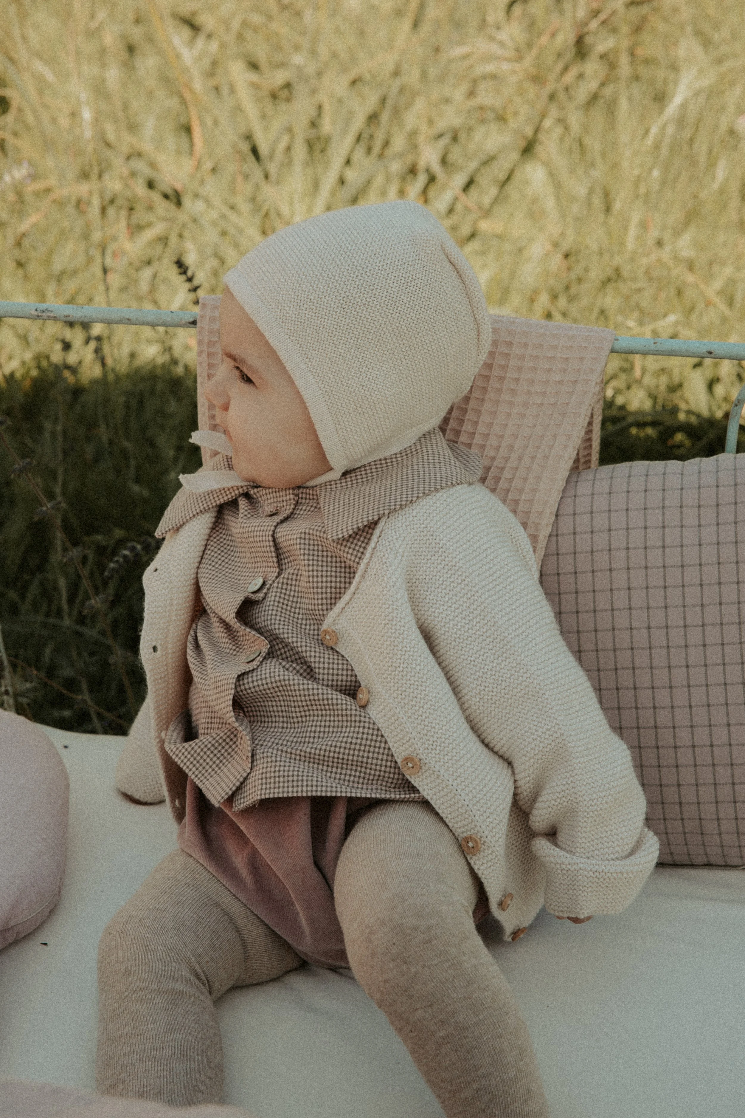 Enfant assis sur un banc, portant une bonnet blanc avec une veste beige et un pantalon rose, dans un environnement naturel.