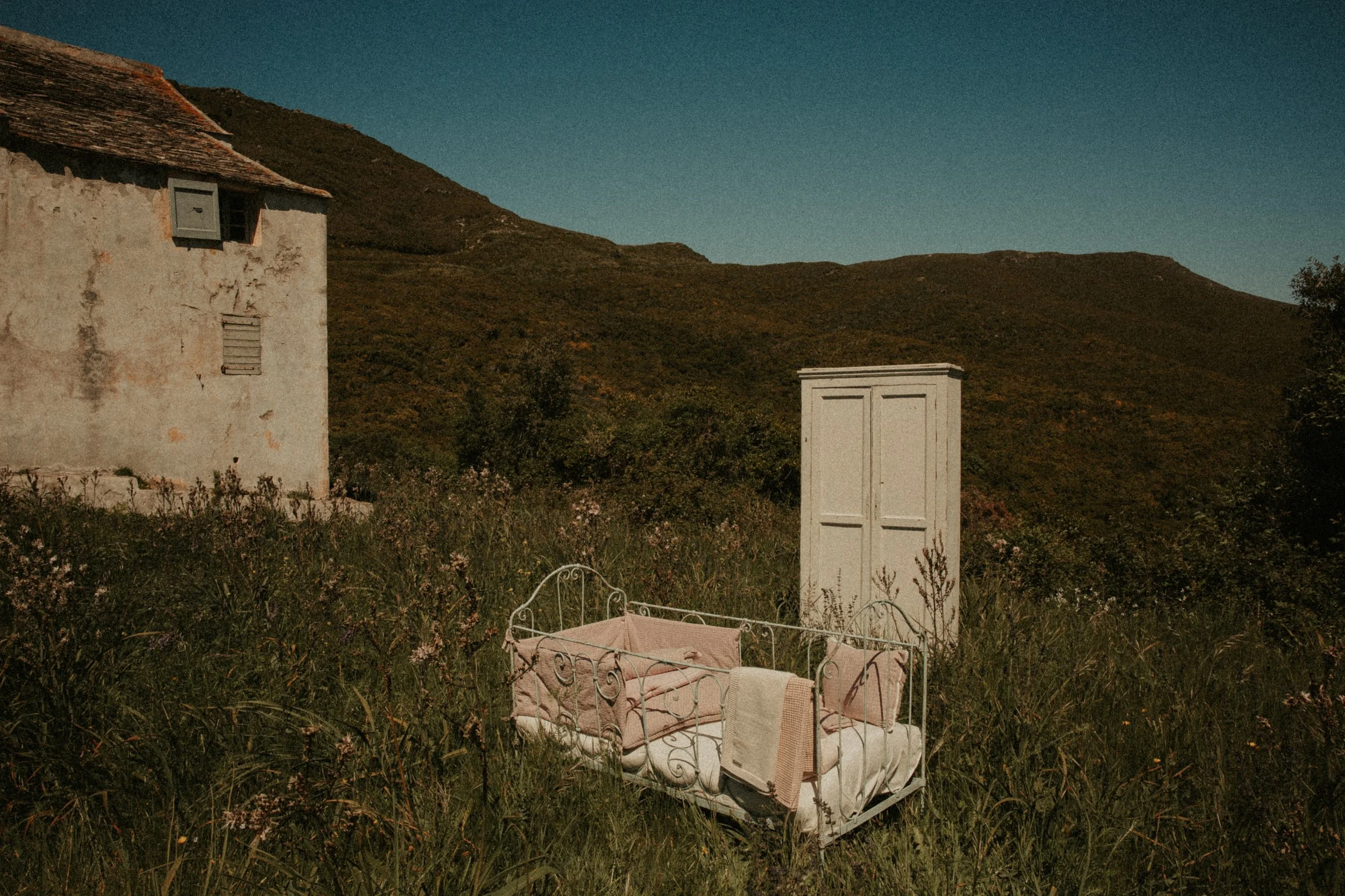 Une cabane ancienne avec un toit en tuiles, située dans un paysage de montagne. Au premier plan, un lit avec des draps et une couverture est placé dans l'herbe, entouré de fleurs et de végétation sauvage.