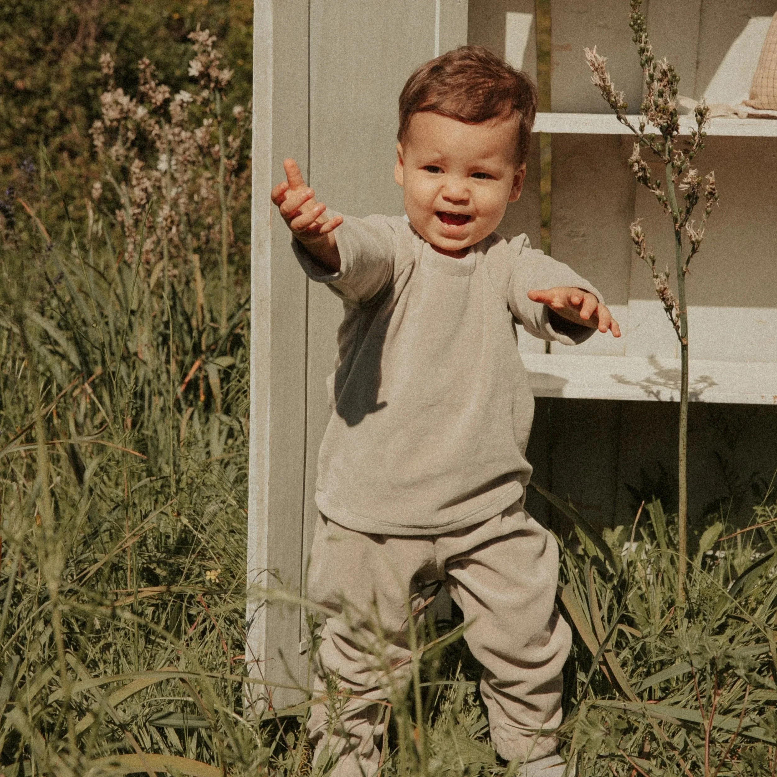 Un jeune garçon avec une chemise beige et un pantalon beige, pointant un doigt vers la caméra, se tenant à côté d'une structure blanche dans un environnement extérieur avec des herbes et des plantes.