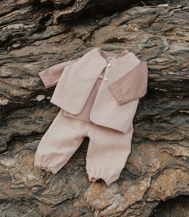 Vêtements pour bébé, comprenant une veste rose pâle avec manches brunes, une chemise rose et un pantalon beige avec rayures, posés sur une surface rocheuse.