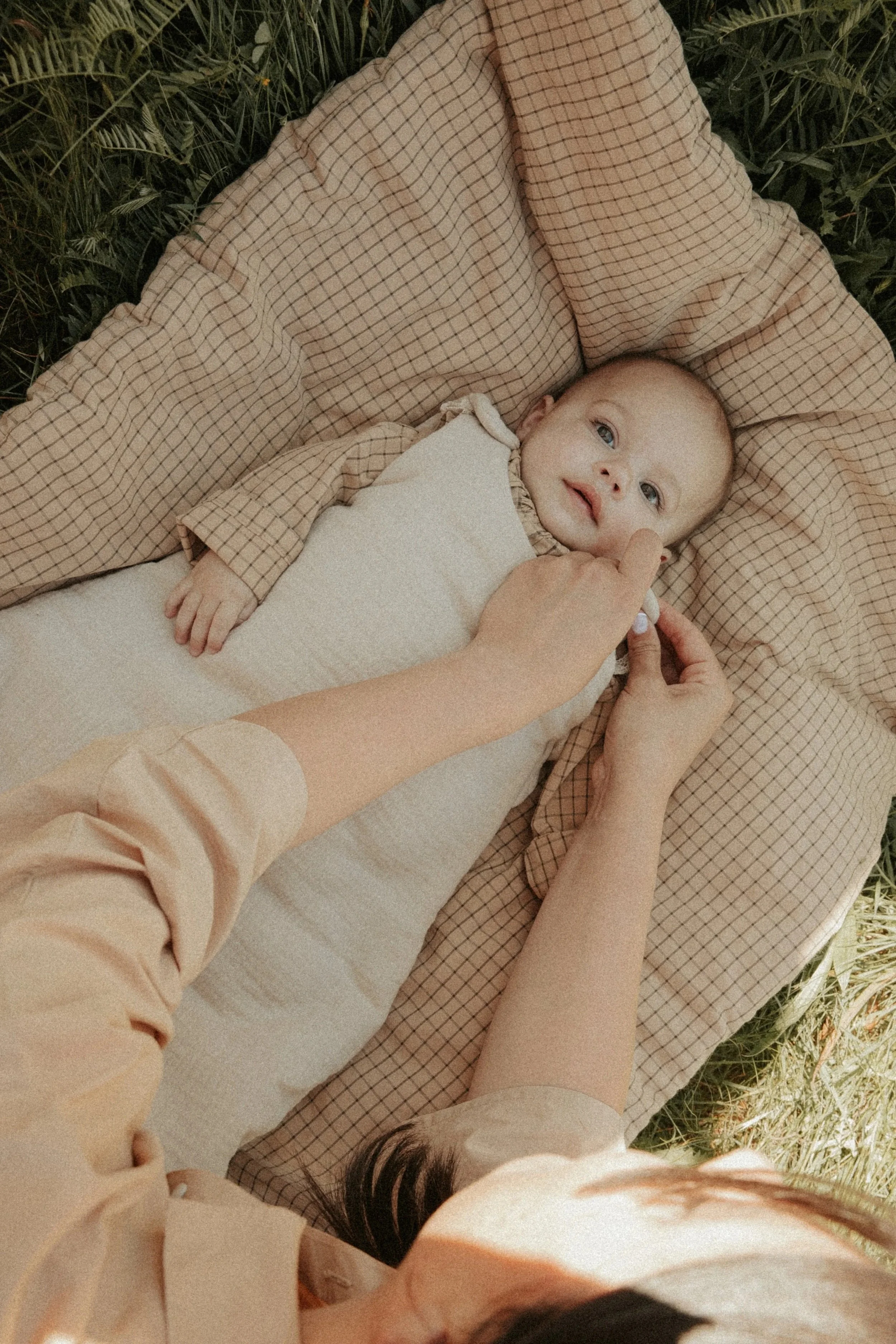 Un bébé allongé sur une couverture dans l'herbe, regardant la caméra, quelques mains ajustent sa position.