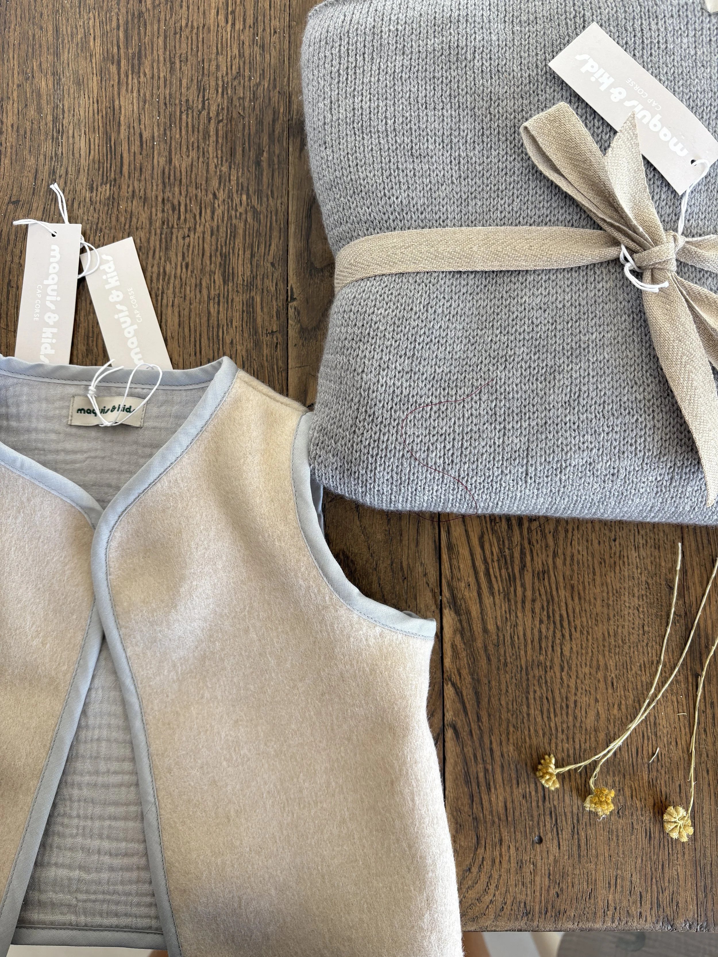 Une nappe en bois sur laquelle sont posés une robe beige en tissu doux, un pull gris en laine et un coussin beige avec une bande beige en tissu, ainsi que quelques fleurs séchées jaunes.