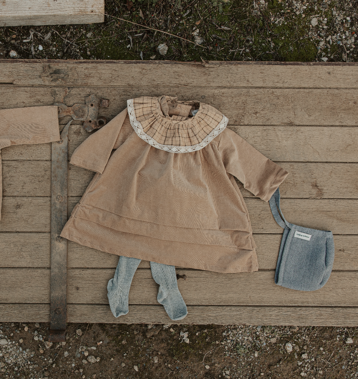 Vêtement pour bébé posé sur un sol en bois, comprenant une robe beige, une paire de chaussettes grises, un chapeau gris et un morceau de tissu beige.