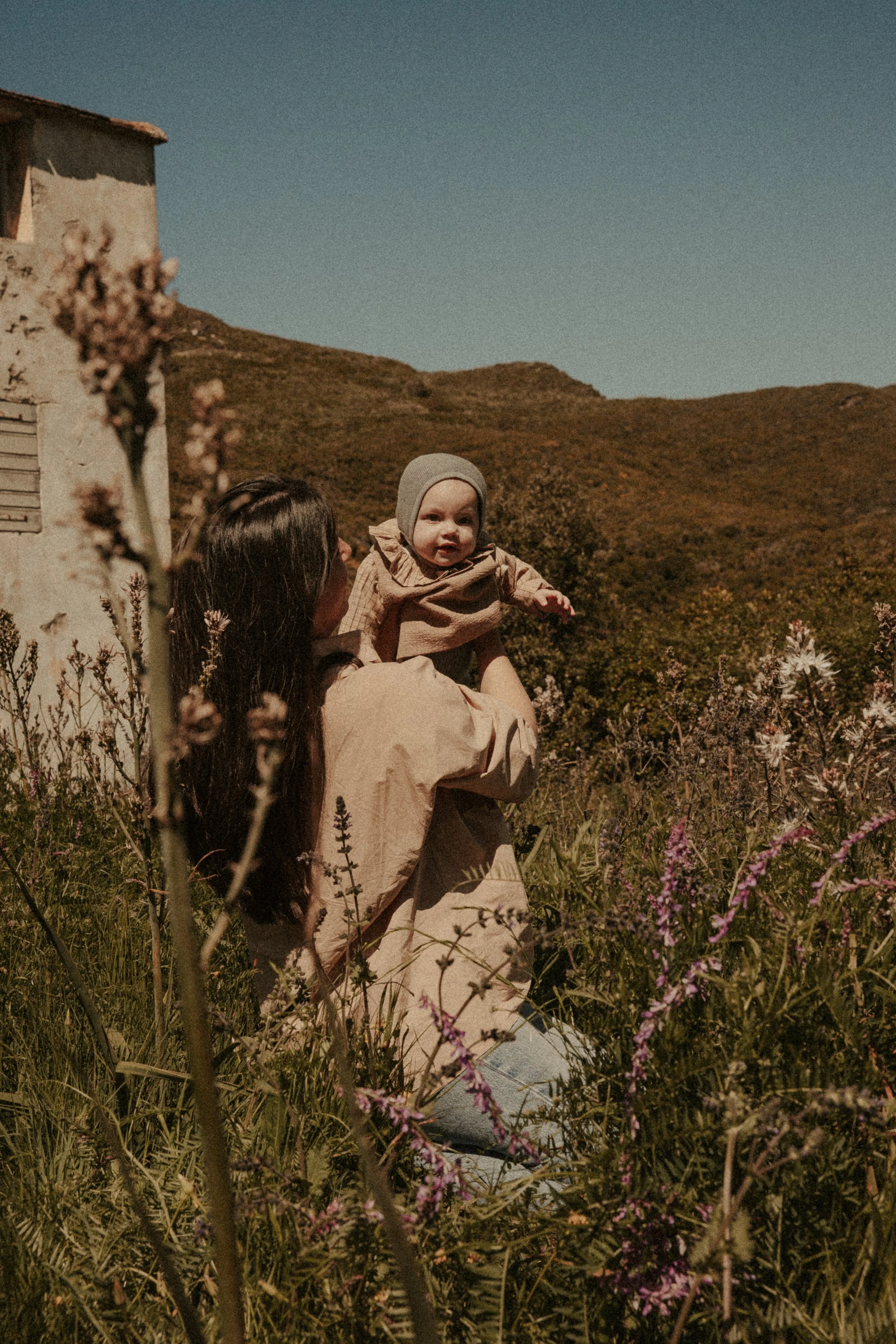 Une femme tient un bébé dans un champ de fleurs, avec des collines en arrière-plan et un ciel clair.