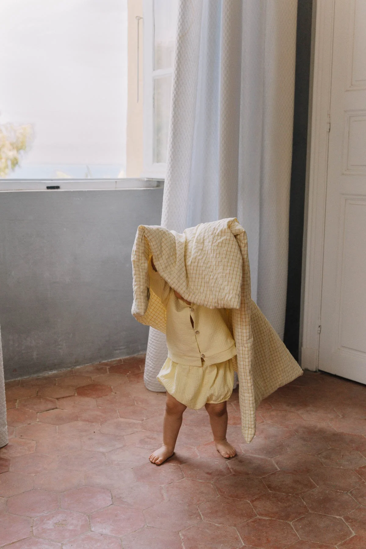 Une personne enfant habillée en jaune avec un vêtement sur la tête, se tenant dans une pièce lumineuse avec des rideaux, un placard blanc et un sol en carrelage rose. La personne cache son visage avec un vêtement.