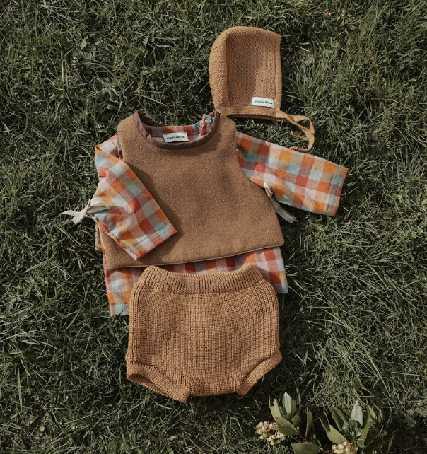Vêtements pour bébé sur de l'herbe, comprenant un haut à manches longues à carreaux orange et blanc, un sous-pull brun, une couche culotte en maille beige, un bonnet en molleton beige, et une capuche assortie.