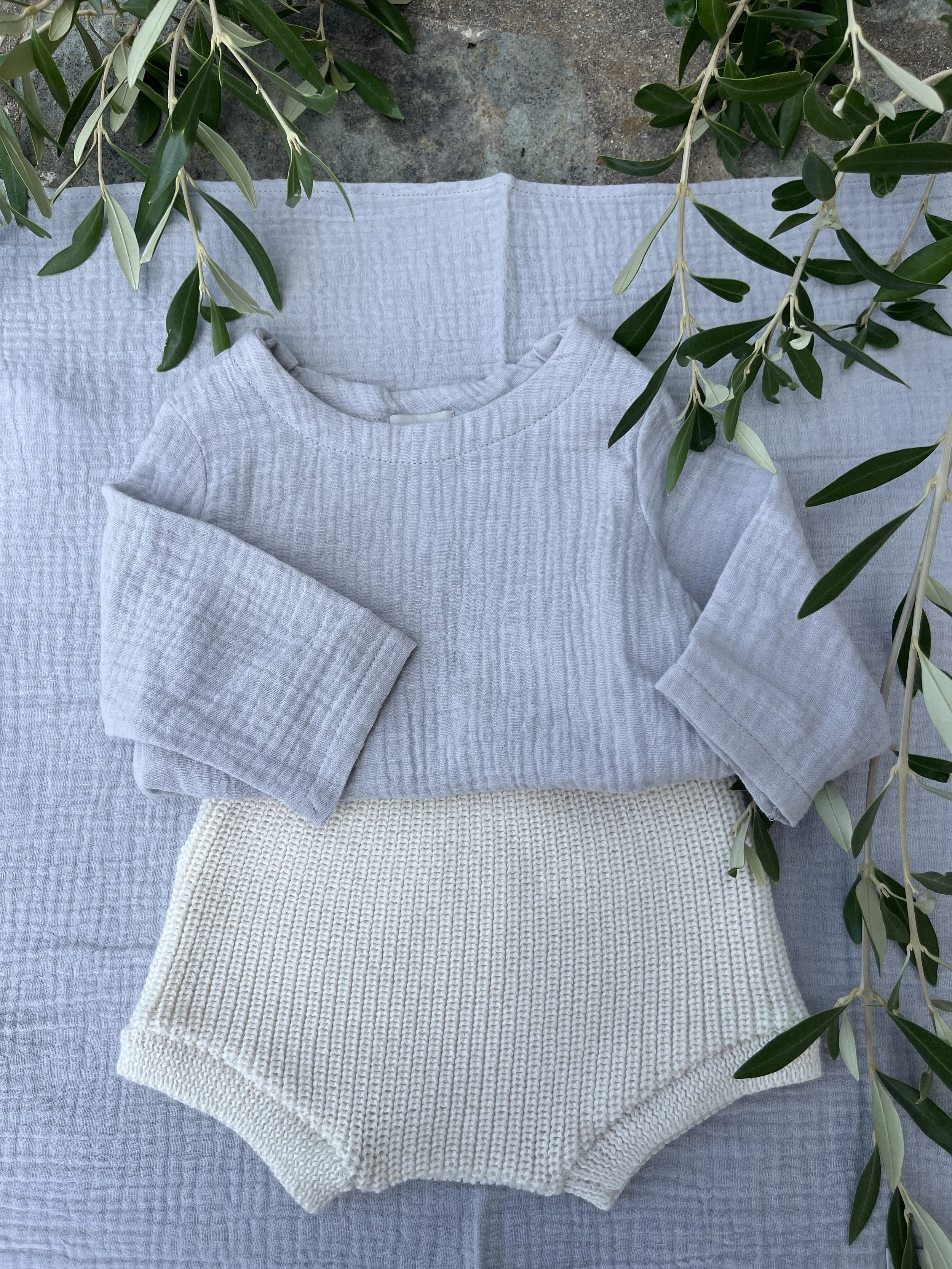 Un vêtement gris en tissu gaufré plié sur un linge de table gris. Il y a aussi un lainage crème plissé et quelques branches d'olivier avec des feuilles vertes.