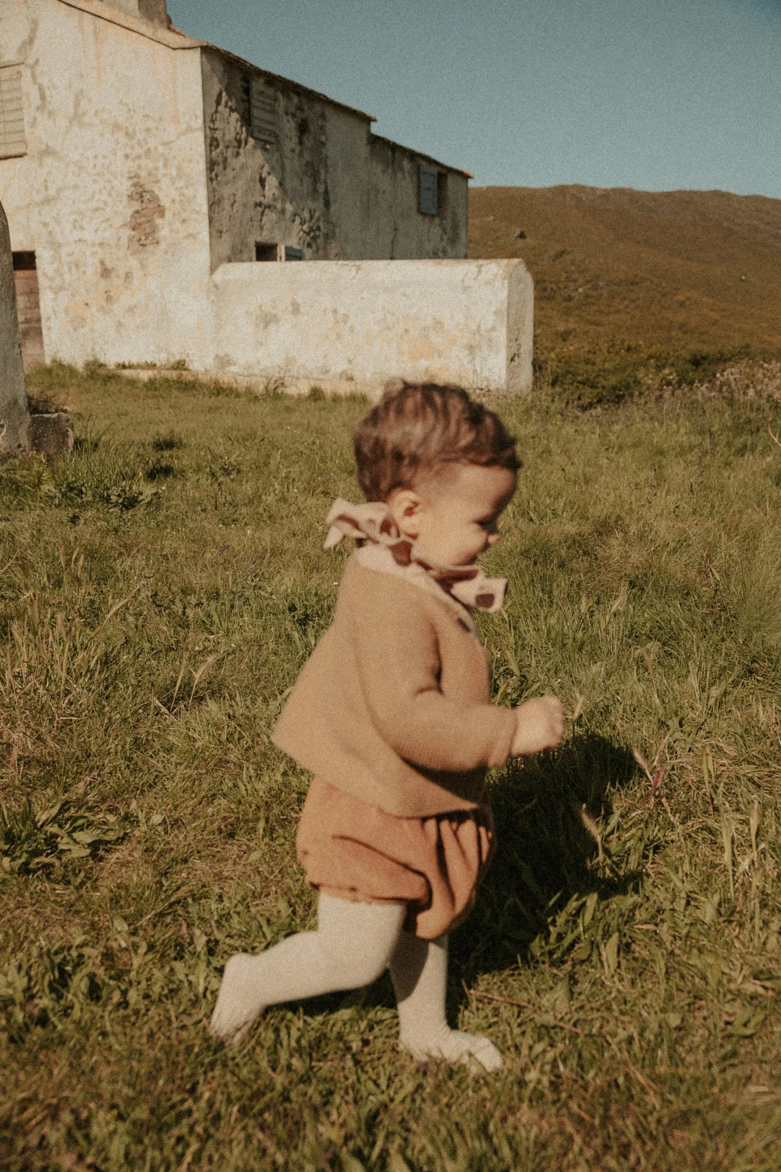 Un jeune enfant marche dans une prairie verte, portant un vêtement beige avec une écharpe à motifs. En arrière-plan, on voit une maison en pierre avec des murs usés et un paysage de collines sous un ciel dégagé.