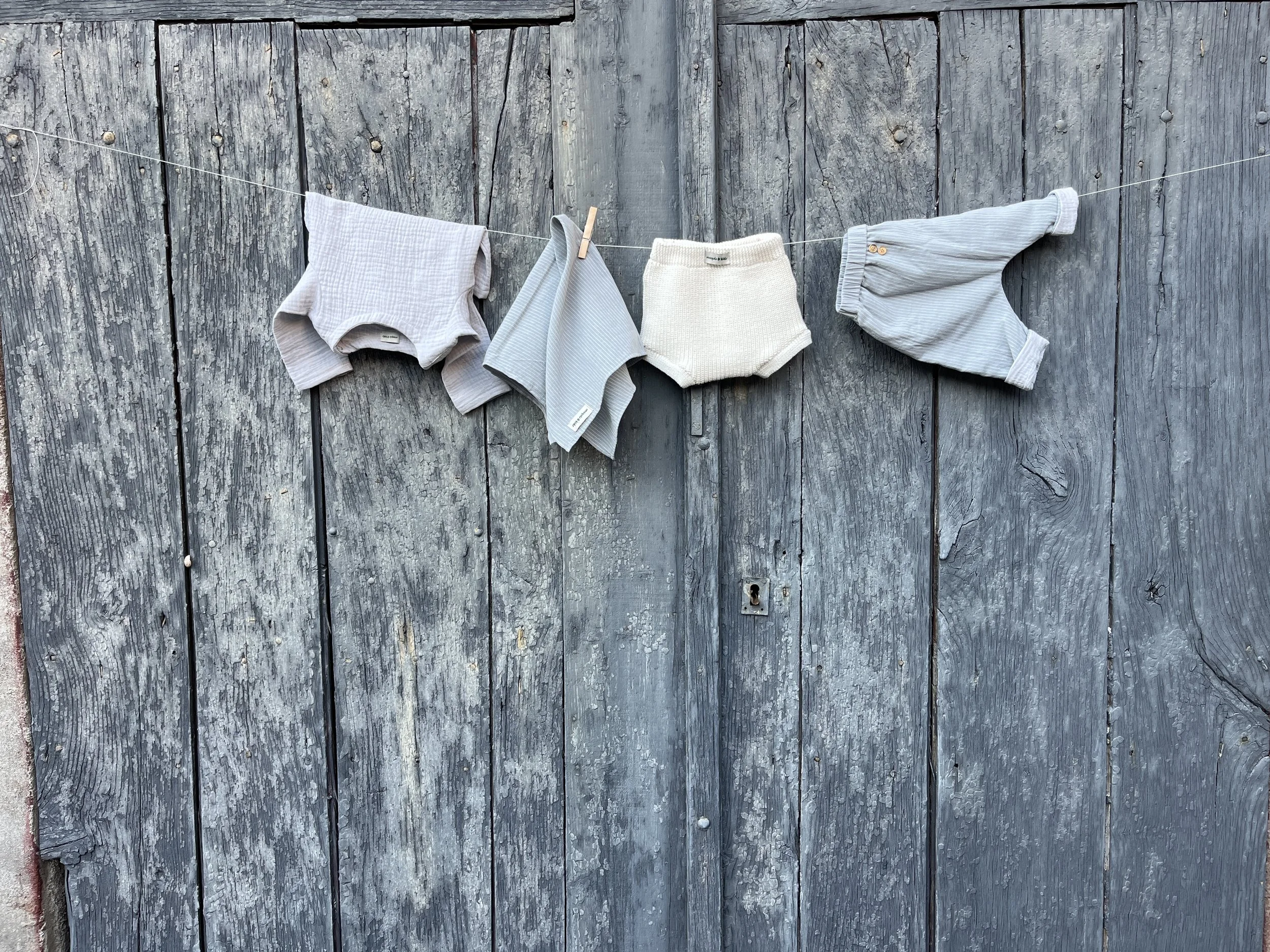 Vêtements pour bébé suspendus sur une corde à linge en bois, contre un vieux mur en bois.