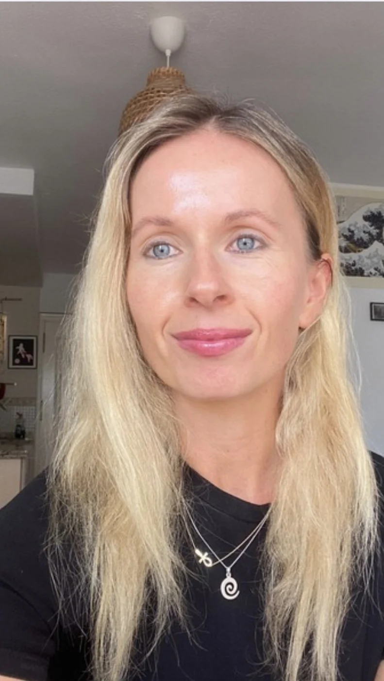 A woman with long blonde hair and blue eyes smiling while wearing a black shirt and layered necklaces, with a kitchen and a light fixture in the background.