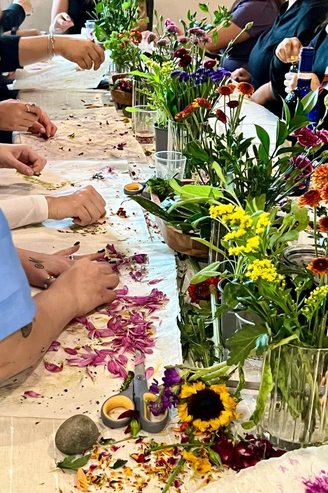 People making floral arrangements with various colorful flowers, petals, scissors, and jars on the table.