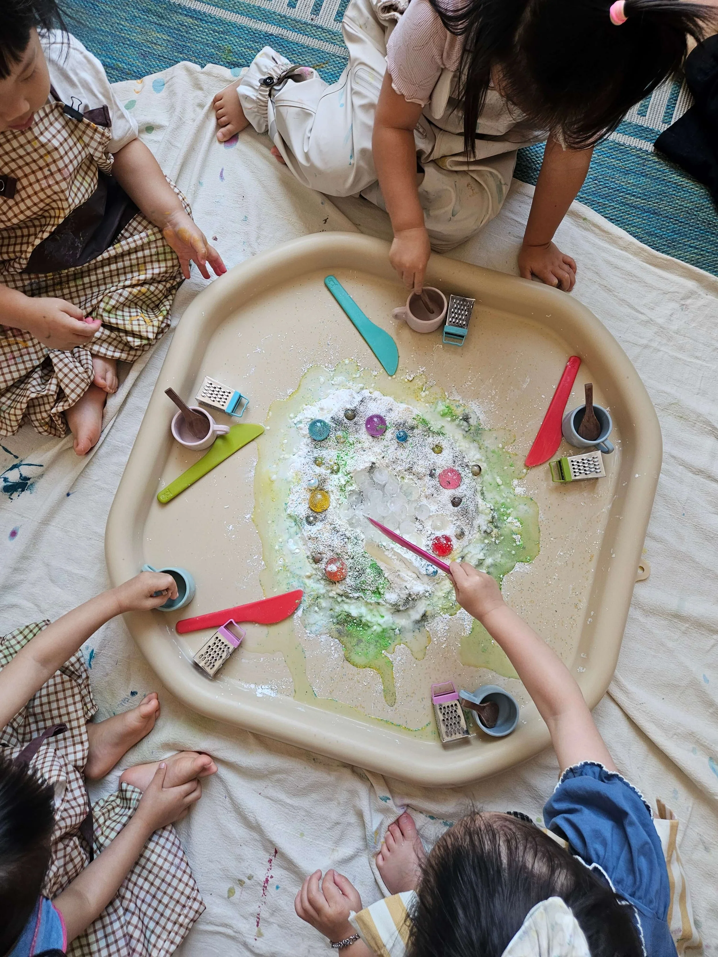 Young children gather around a sensory tray to scoop, mix, and explore materials together at Whimsy Art Club.