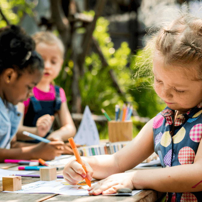 Group of children drawing and coloring outdoors at a wooden table with colorful markers and paper.