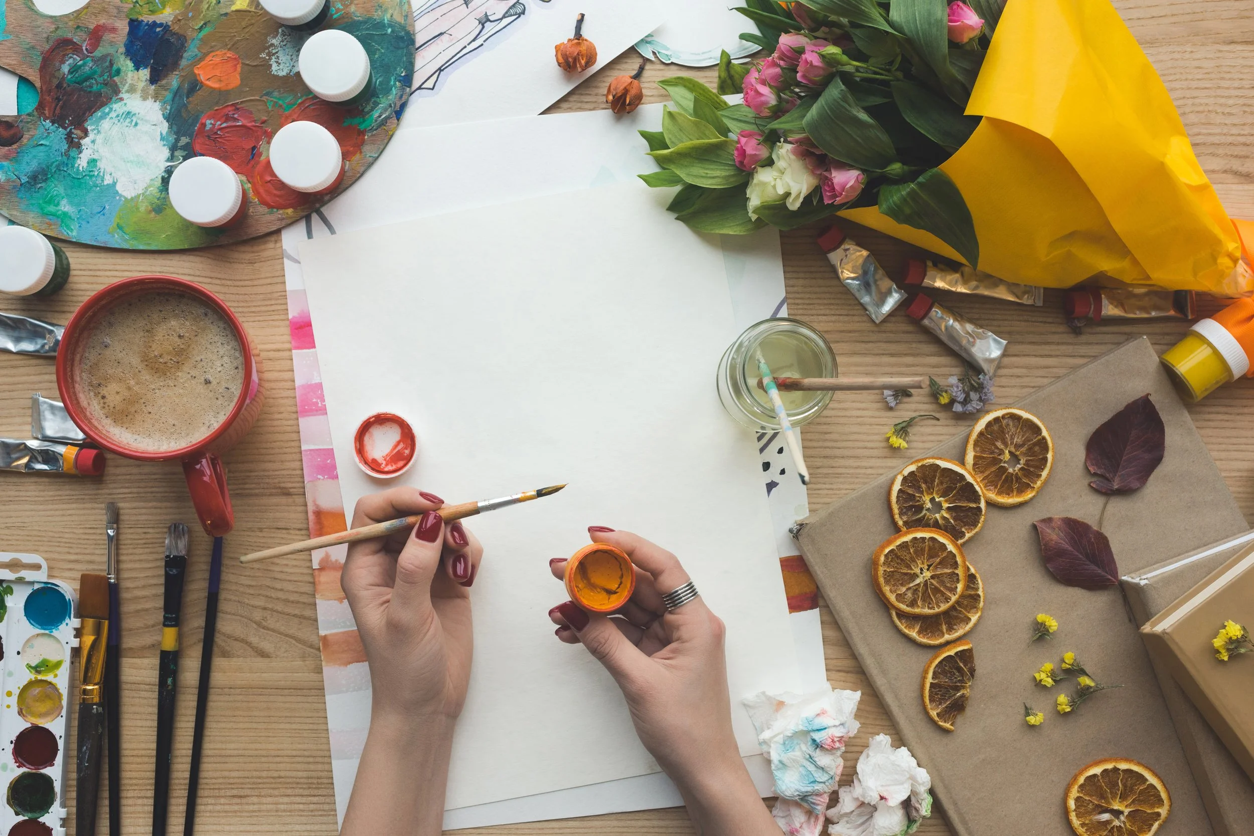 Artist painting with watercolors on blank paper, surrounded by art supplies, dried orange slices, and flowers on a wooden table.
