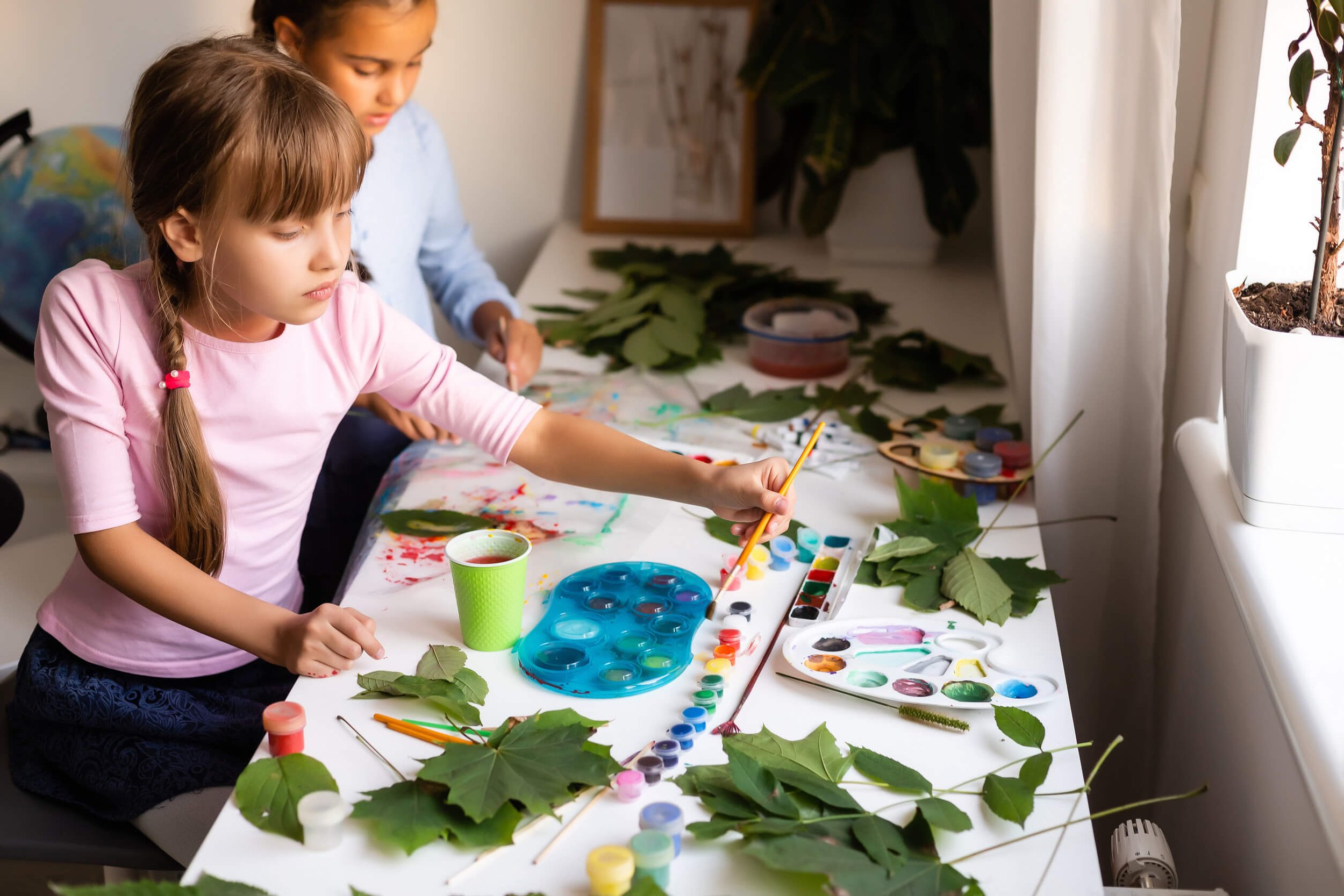 Two young girls are painting and crafting with leaves and nature-inspired materials at a table in a well-lit room.