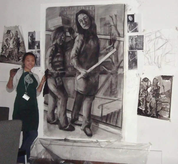 A young girl standing and smiling next to a large black and white painting of two women in an indoor art studio, surrounded by smaller artworks on the wall.