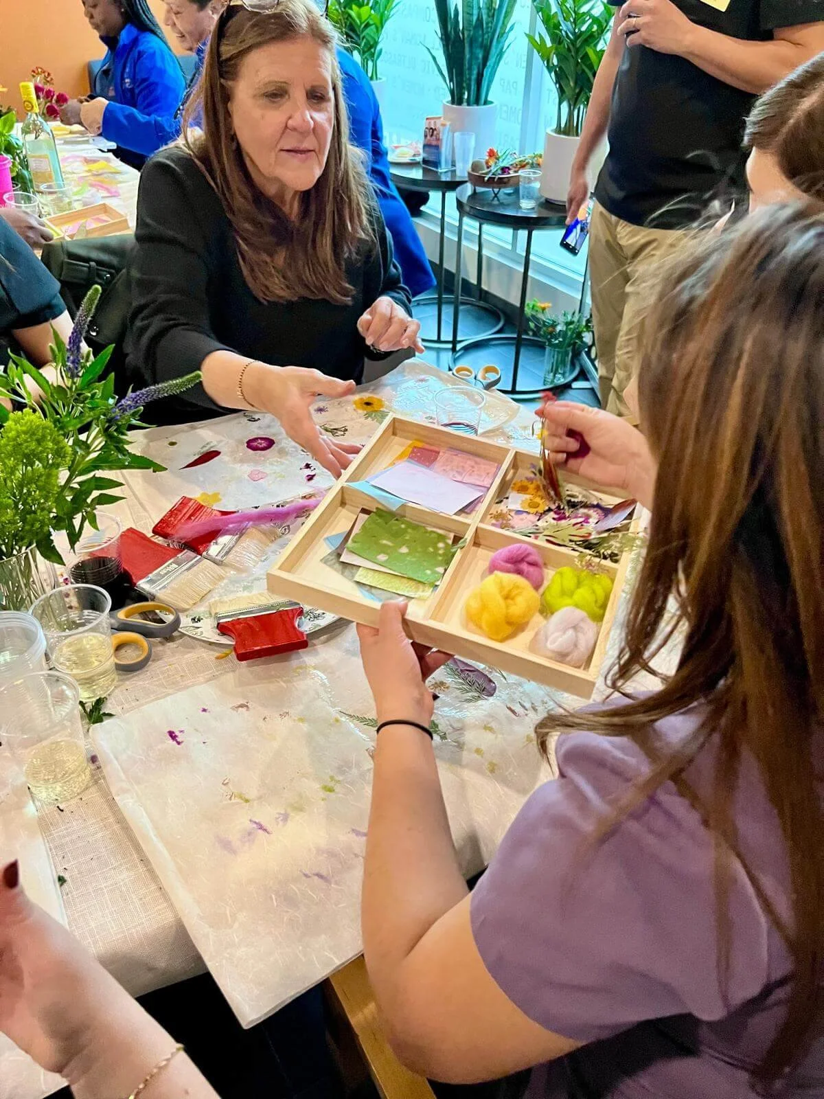 A group of people gathered at a table participating in a crafting activity with flowers and colorful felted balls, with a woman in black receiving supplies from another woman in a light purple shirt.