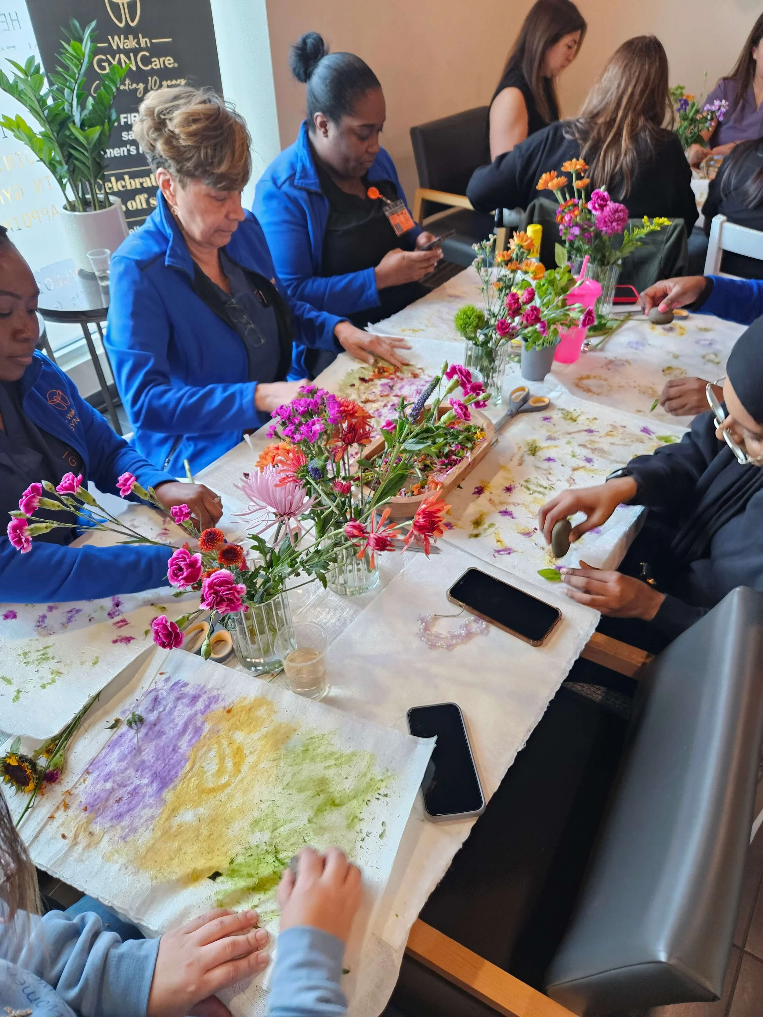 Group of women participating in a flower arranging workshop at a long table. The table is decorated with colorful flowers, scissors, and materials for flower arranging.
