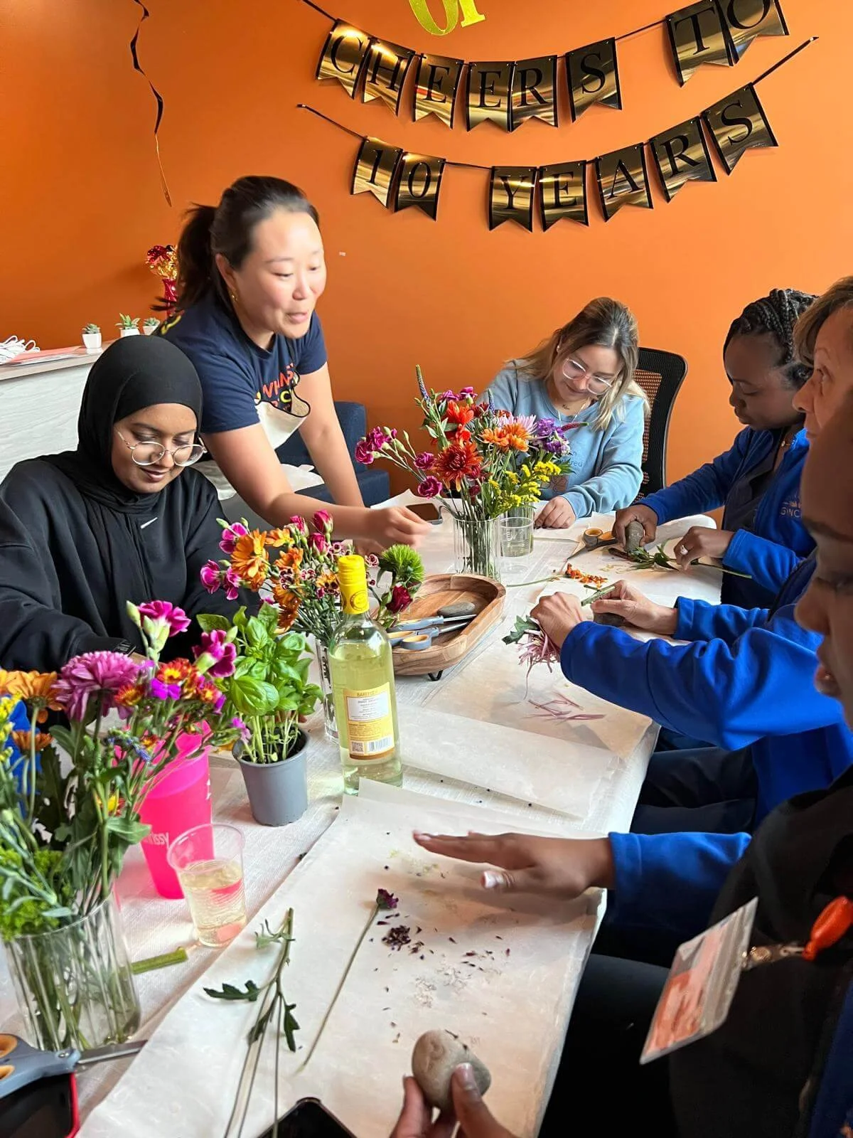 Group of women and girls gathered around a table, making floral arrangements for a celebration, with colorful flowers, tools, and a 'Cheers to 10 Years' banner in the background.