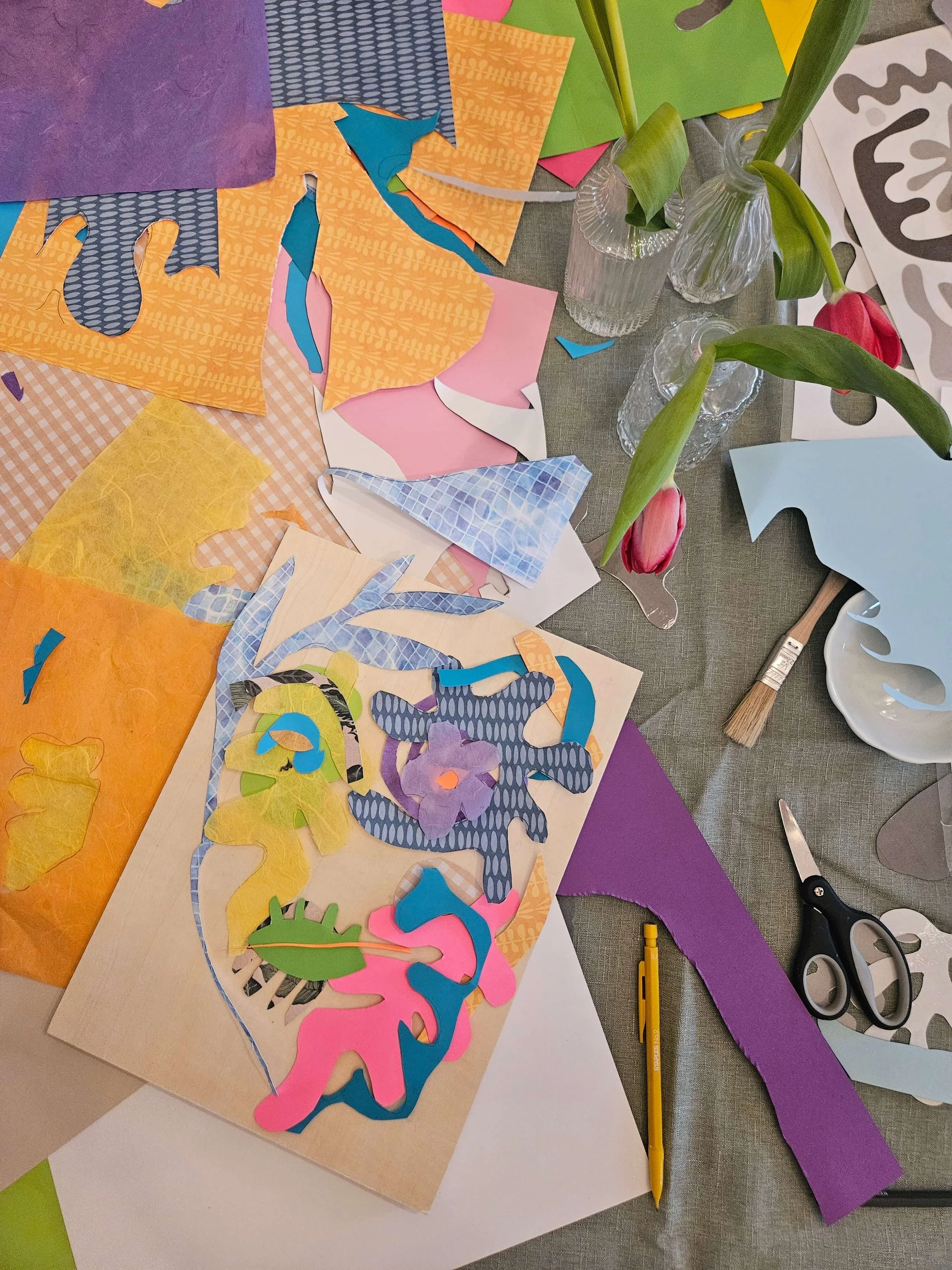 Colorful paper cutouts and crafting supplies on a table, with pink tulips in glasses and various patterned papers.