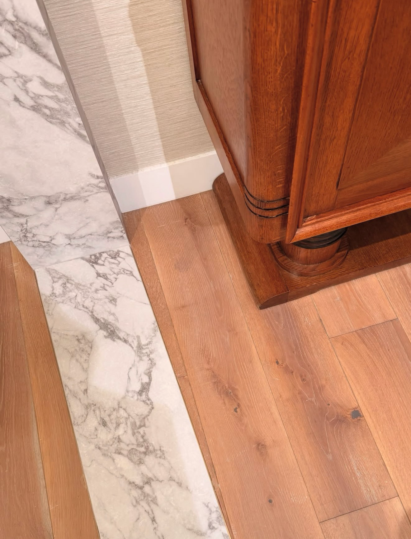 Close-up of a decorative hardwood floor with a marble tile border next to a wooden cabinet and baseboard, showing detailed wood grain and marble veining.