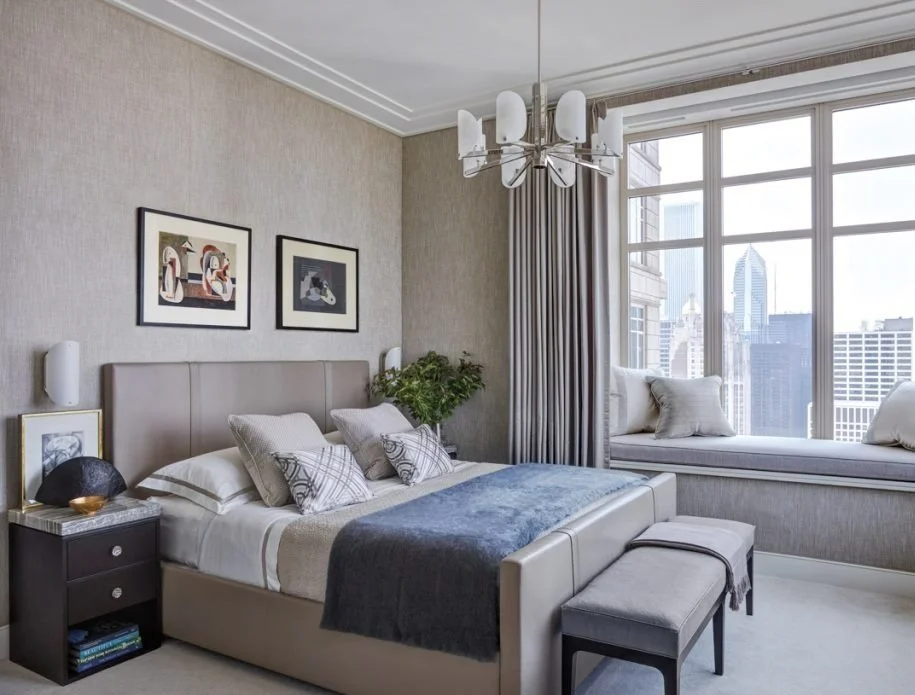 Modern bedroom with neutral tones, artwork on the wall, a bed with decorative pillows and a dark throw blanket, a nightstand with decorative objects, a window seat with cushions by large windows showing city skyscrapers outside, and a contemporary ch