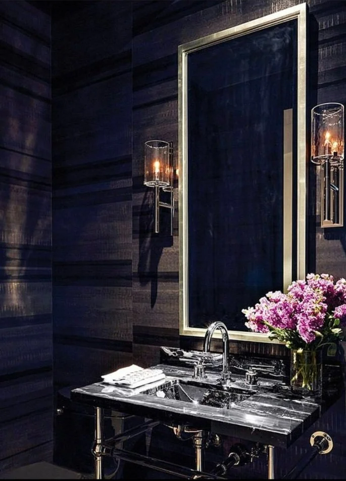 A luxurious bathroom with a black marble sink, a large framed mirror, dark wooden wall panels, and two modern wall sconces with glass covers and lit candles. There is a bouquet of pink flowers on the right side of the sink, and neatly folded white to