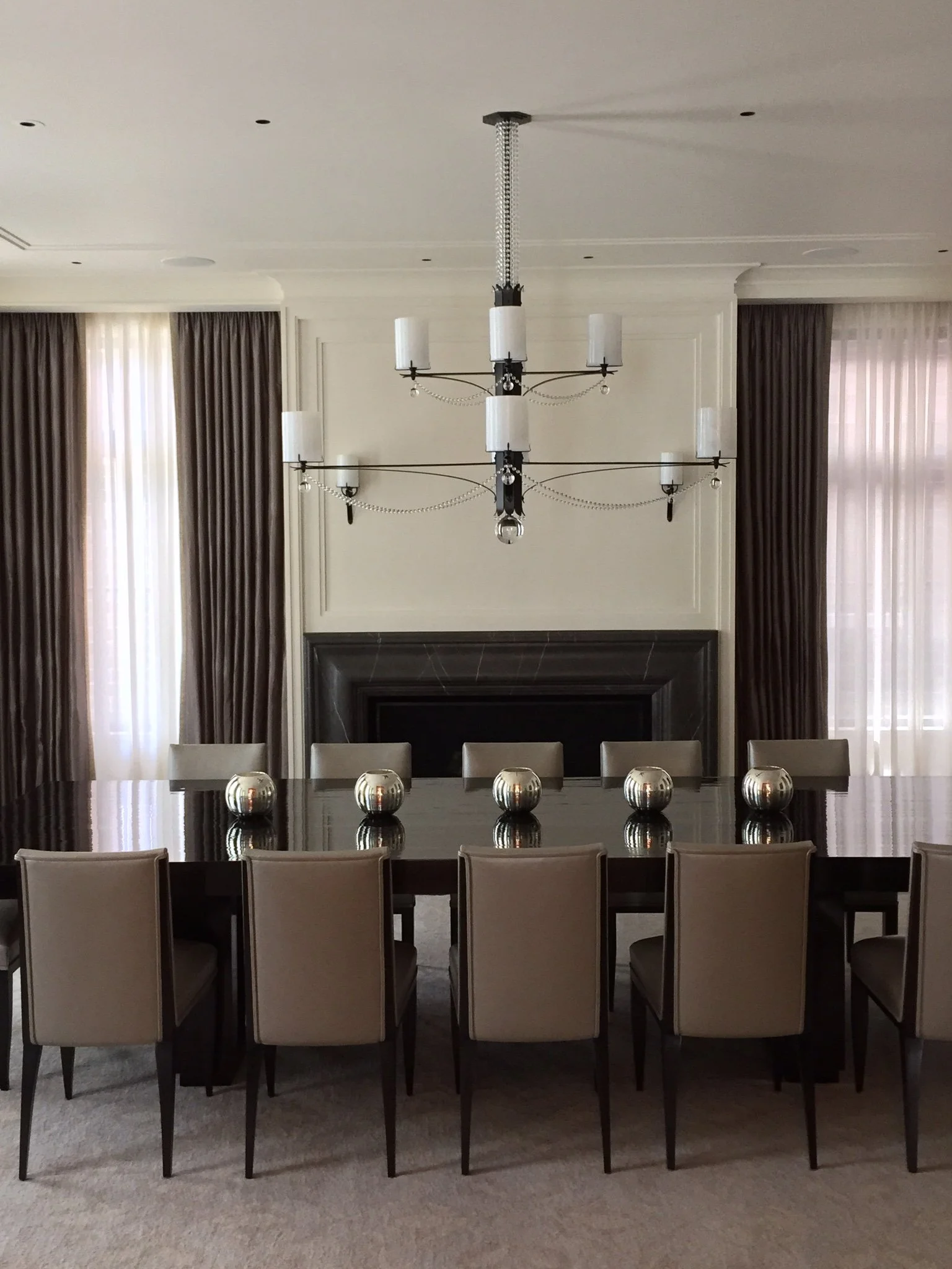 A formal dining room with a large dark wood table surrounded by beige chairs, a modern black chandelier with multiple candle-like lights, and a fireplace with beige curtains on either side.