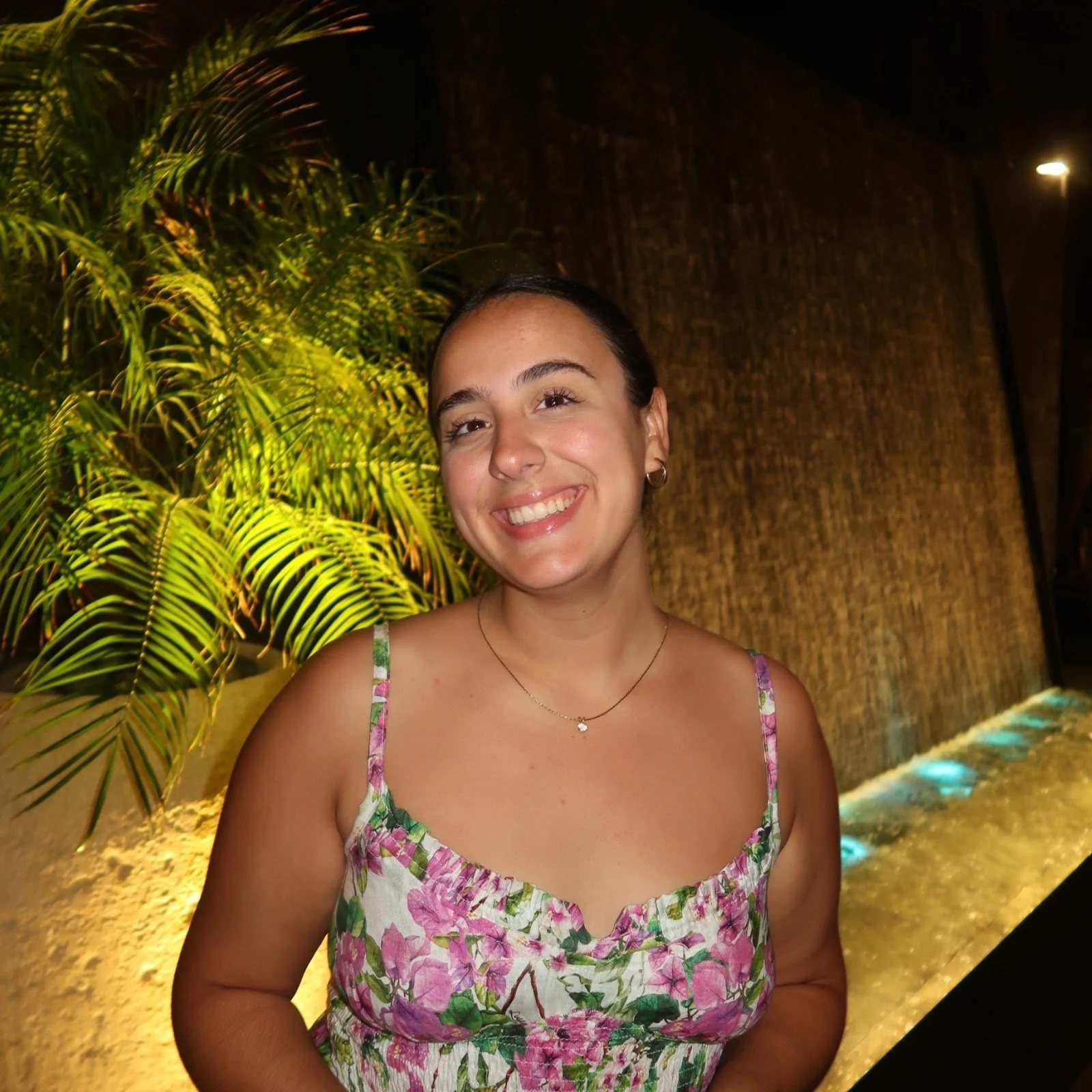 Smiling woman in floral dress beside illuminated waterfall and plants at night.