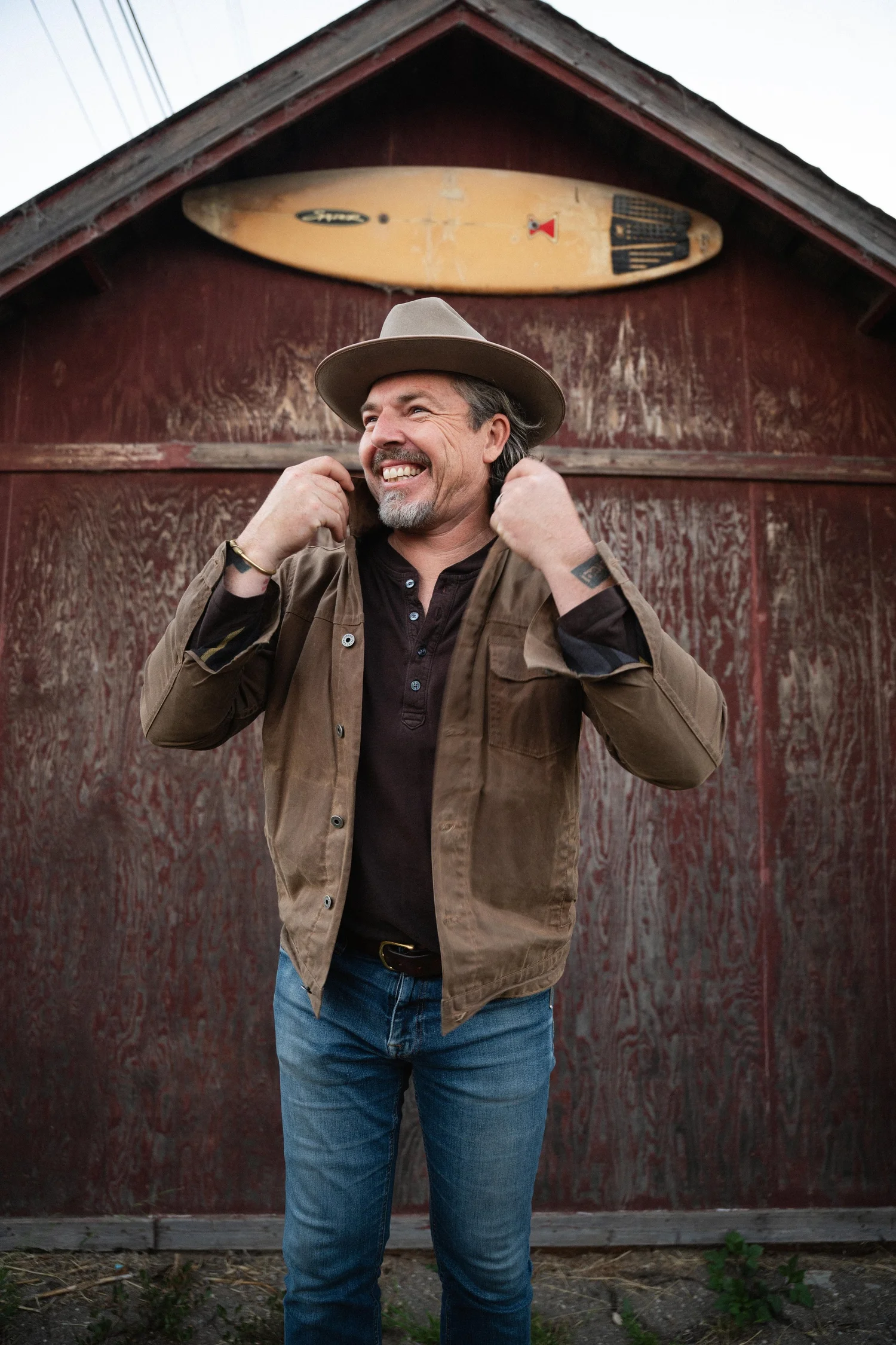 Neil Broere smiling in a wide-brimmed hat in front of a red barn wall with a surfboard